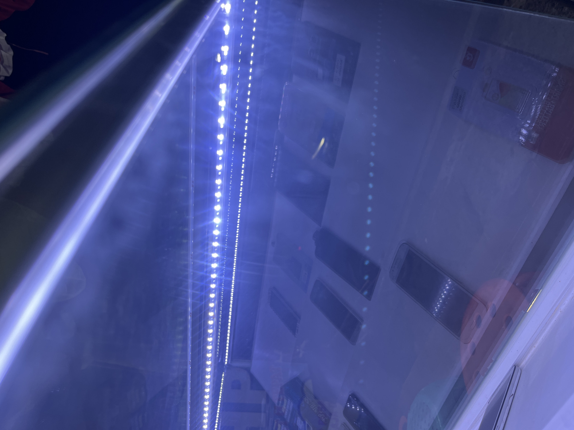 The image shows a display of smartphones under bright blue LED lights, likely in a retail store. The phones are arranged on a white surface behind a glass enclosure. Several phones are visible, some with their screens on, others off. The lighting creates reflections on the glass and the surfaces of the phones. In the background, there's a hint of merchandise or signage. The overall impression is of a modern electronics display. The image does not contain any information that would allow for the determination of its location in the city of Central Jakarta, Indonesia.