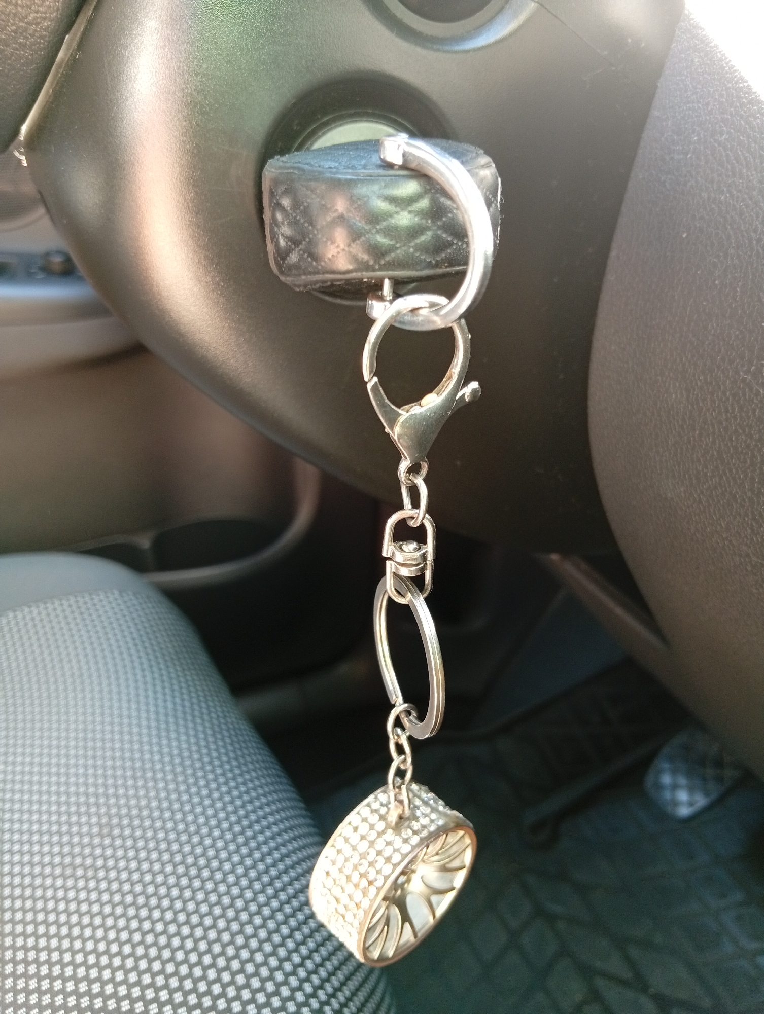 A close-up shot shows a car's ignition with a key in it, attached to a keychain. The keychain is decorated with a diamond-like charm and a leather fob. The background features part of a car's interior, including the steering wheel and a seat. The image is taken from a slightly low angle, focusing on the ignition and keychain.