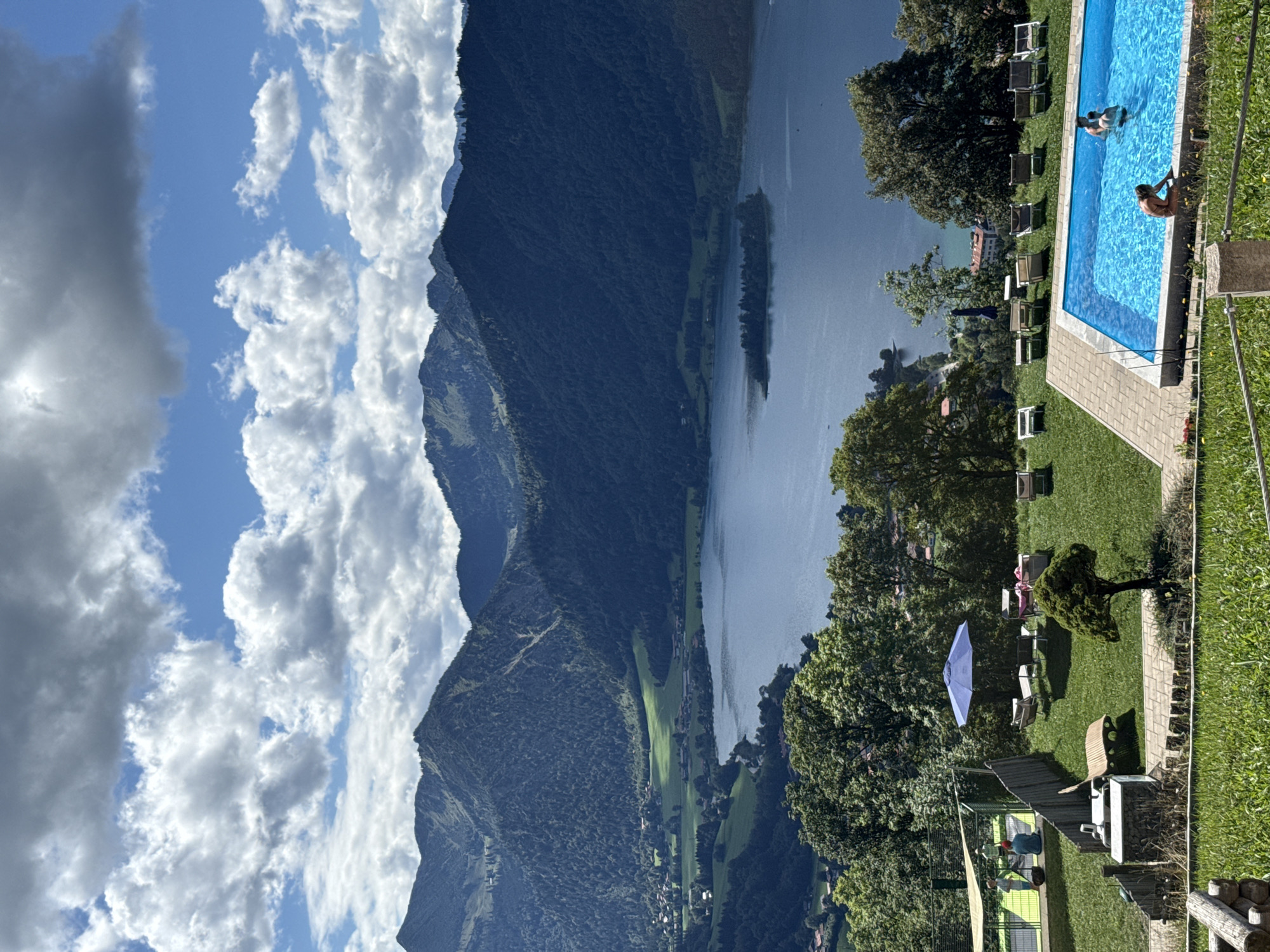 This is a high-angle, medium shot of a resort area in Schliersee, Germany, with a swimming pool in the foreground and a lake and mountains in the background. The sky is partly cloudy with dramatic white and gray clouds. The mountains are covered in dark green evergreen trees. The lake is calm and blue.
In the foreground, a rectangular swimming pool filled with bright blue water is the central focus. Three people are visible in the pool; two are partially submerged and appear to be interacting, while a third person sits on the edge of the pool. Around the pool, lounge chairs and a blue umbrella are set up on a well-maintained green lawn. To the left of the pool, a tennis court with a green surface is visible, with two people standing near the net.
Further in the background, a densely wooded hillside slopes down towards the lake. Small buildings and scattered trees are visible on the slopes and near the lake's edge. The overall scene conveys a sense of leisure and natural beauty.