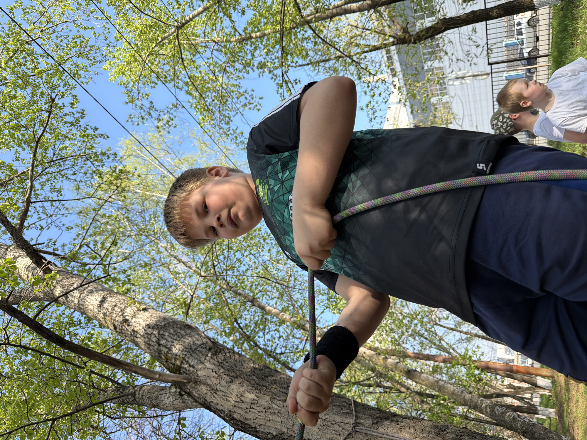 The image shows a young boy, likely around 10-12 years old, holding a climbing rope. He is wearing a dark t-shirt with a green geometric pattern, dark blue pants, and a black wristband on his left wrist. He is looking down at the rope, which is running through his hands. Behind him, out of focus, a younger boy can be seen wearing a white t-shirt and looking up. The background consists of trees with fresh green leaves and a clear blue sky. Also visible are the wires and building. Based on visual clues, it seems the image was taken in a park or green area, possibly in Izhevsk, Russia.
