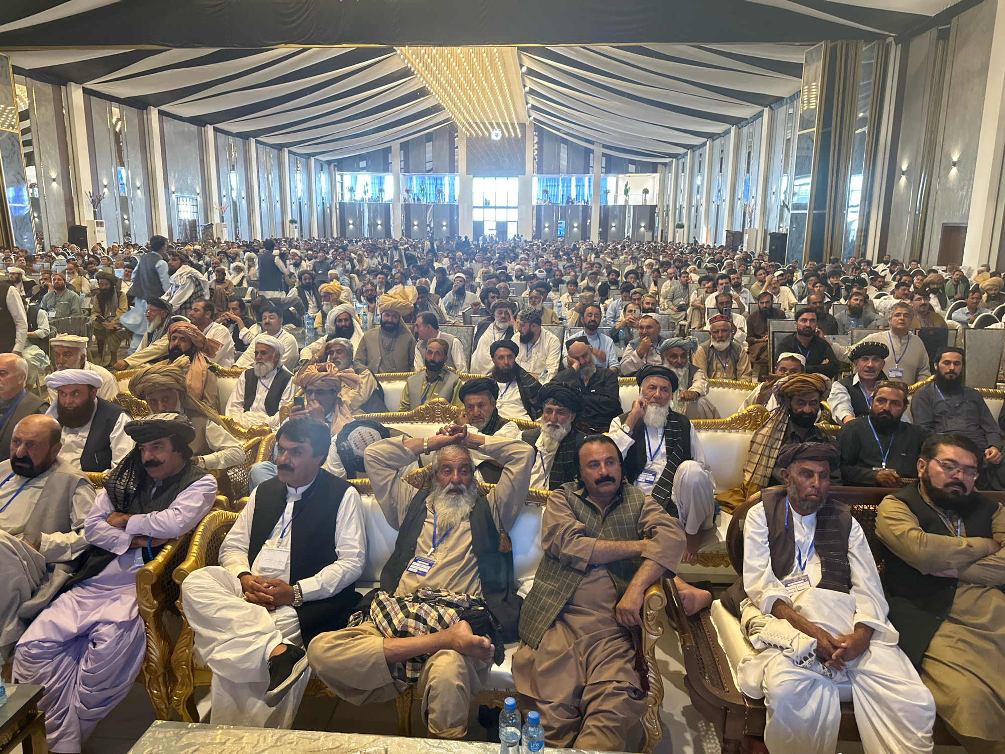 The image captures a large gathering of men inside a spacious, ornately decorated hall in Quetta City Tehsil, Pakistan. The venue features a high ceiling with dramatic black and white striped fabric draping, interspersed with reflective panels and decorative lighting fixtures. The attendees are seated on rows of plush, golden-colored chairs, many wearing traditional attire, including turbans and vests.
The men, predominantly of adult age, are engaged in conversation, listening, or observing the proceedings. Some are wearing lanyards with identification badges, suggesting an organized event. The atmosphere appears to be formal, possibly a conference, political rally, or religious gathering. The backdrop suggests a modern, large-scale indoor facility. The lighting is bright, illuminating the crowd and the elaborate ceiling decorations. Several water bottles are visible on a table in the foreground, indicating the duration and nature of the event. The overall scene depicts a significant assembly of people in a public gathering space within Quetta City Tehsil.