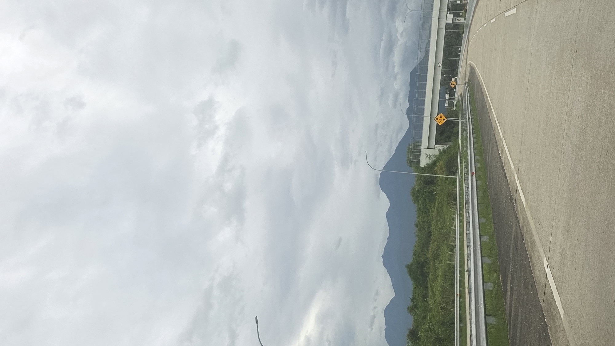 The image shows a highway in Lampanah Tunong, Indonesia. The road is paved and has a white line marking the edge. A metal guardrail separates the road from a green area with trees and bushes. In the background, there are mountains partially obscured by clouds. The sky is overcast with gray clouds. A road sign is visible on the right side of the road. An overpass structure is also visible in the background. There are no people in the frame.