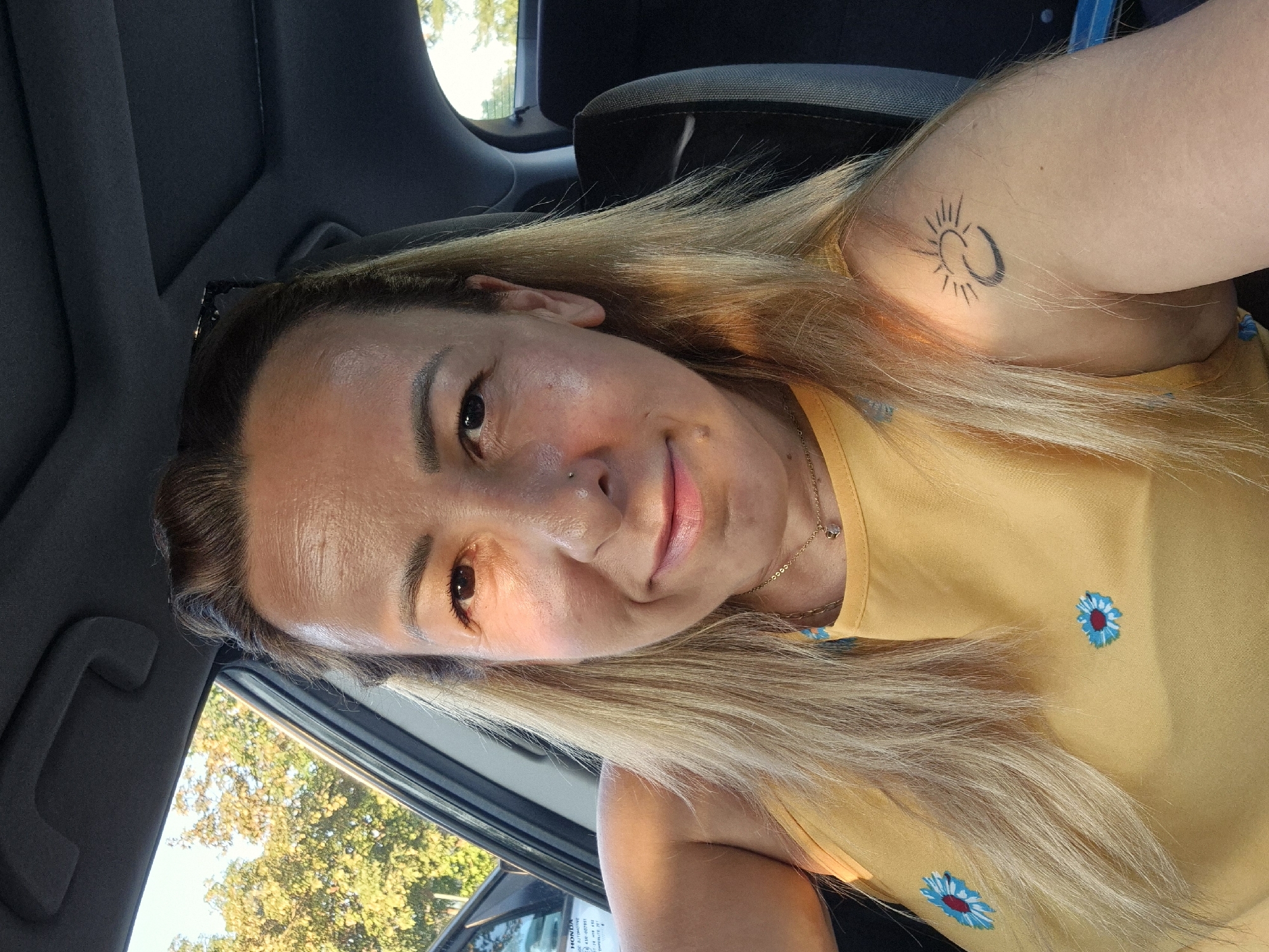 A woman with blonde hair and a yellow tank top with embroidered blue and red flowers sits in the driver's seat of a car. She has a tattoo of a sun and moon on her left shoulder. She is smiling at the camera, and the sun is shining through the car window, casting a warm glow on her face. Outside the car, green trees are visible.