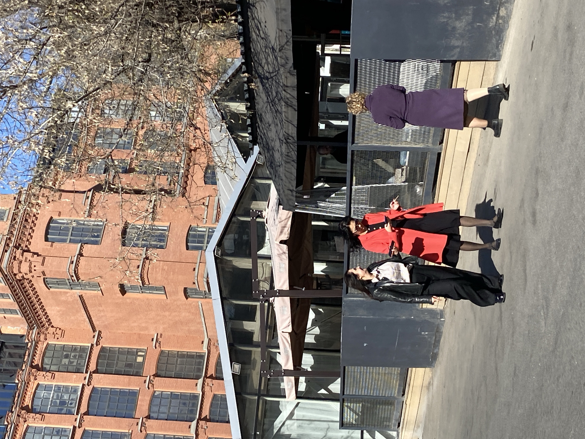 The image shows a brick building in Moscow, Russia, near a cafe. Three people are walking on the sidewalk, one in a purple dress, one in a red coat, and one in a black jacket.