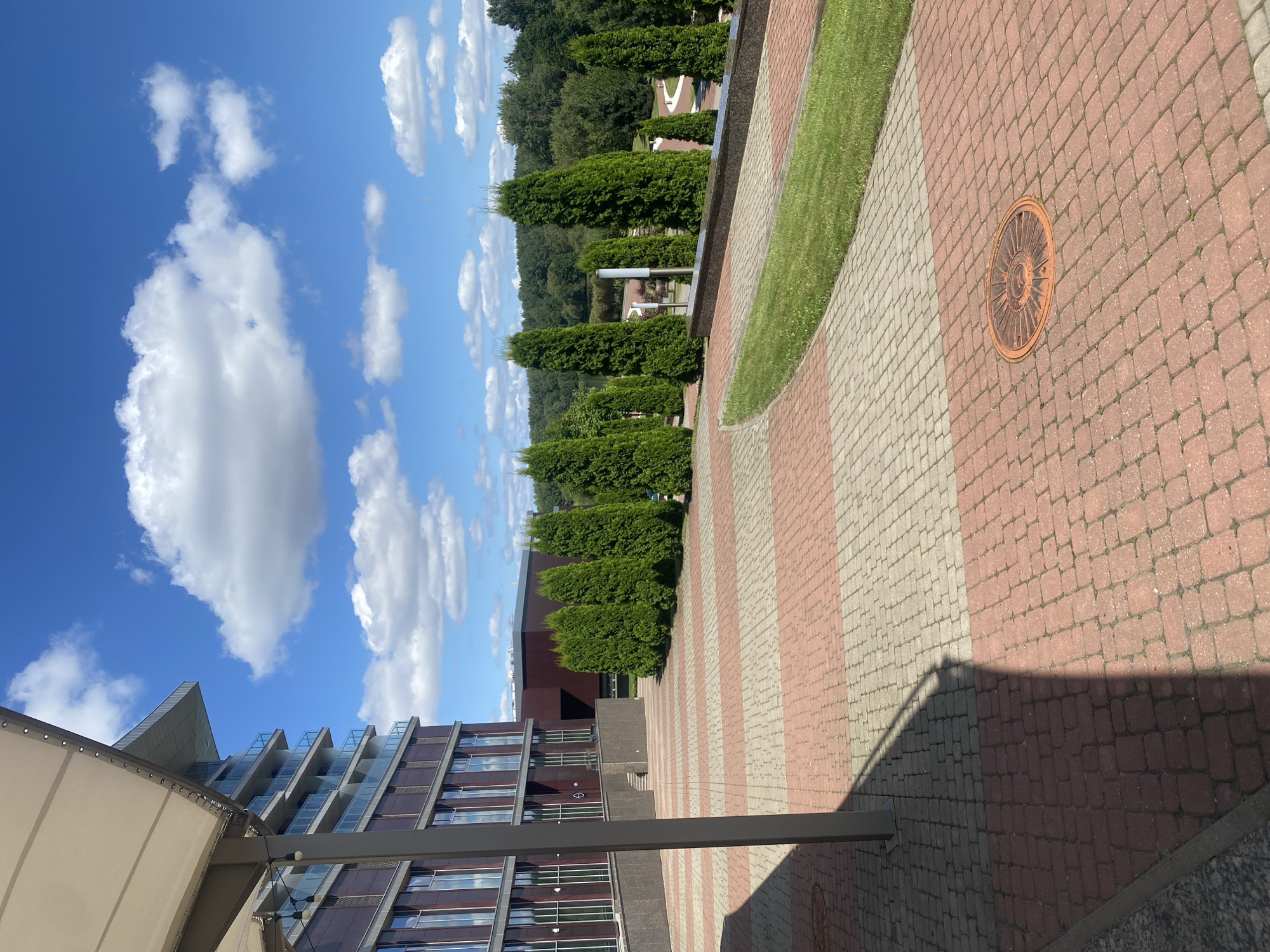 This is an outdoor, eye-level, daytime, medium-shot photograph taken in the city of Minsk, Belarus. The scene depicts a modern building with a glass facade and balconies, adjacent to a paved area with a pattern of red and beige rectangular stones. Several tall, narrow green trees are planted in a line along a raised concrete border. Beyond the trees, a lush green landscape with more trees and a body of water can be seen under a bright blue sky with white, fluffy clouds. A circular orange manhole cover is visible in the foreground on the paved area. A tall, slender grey pole casts a long shadow across the pavement. The overall impression is one of a well-maintained, contemporary urban environment integrated with nature.
