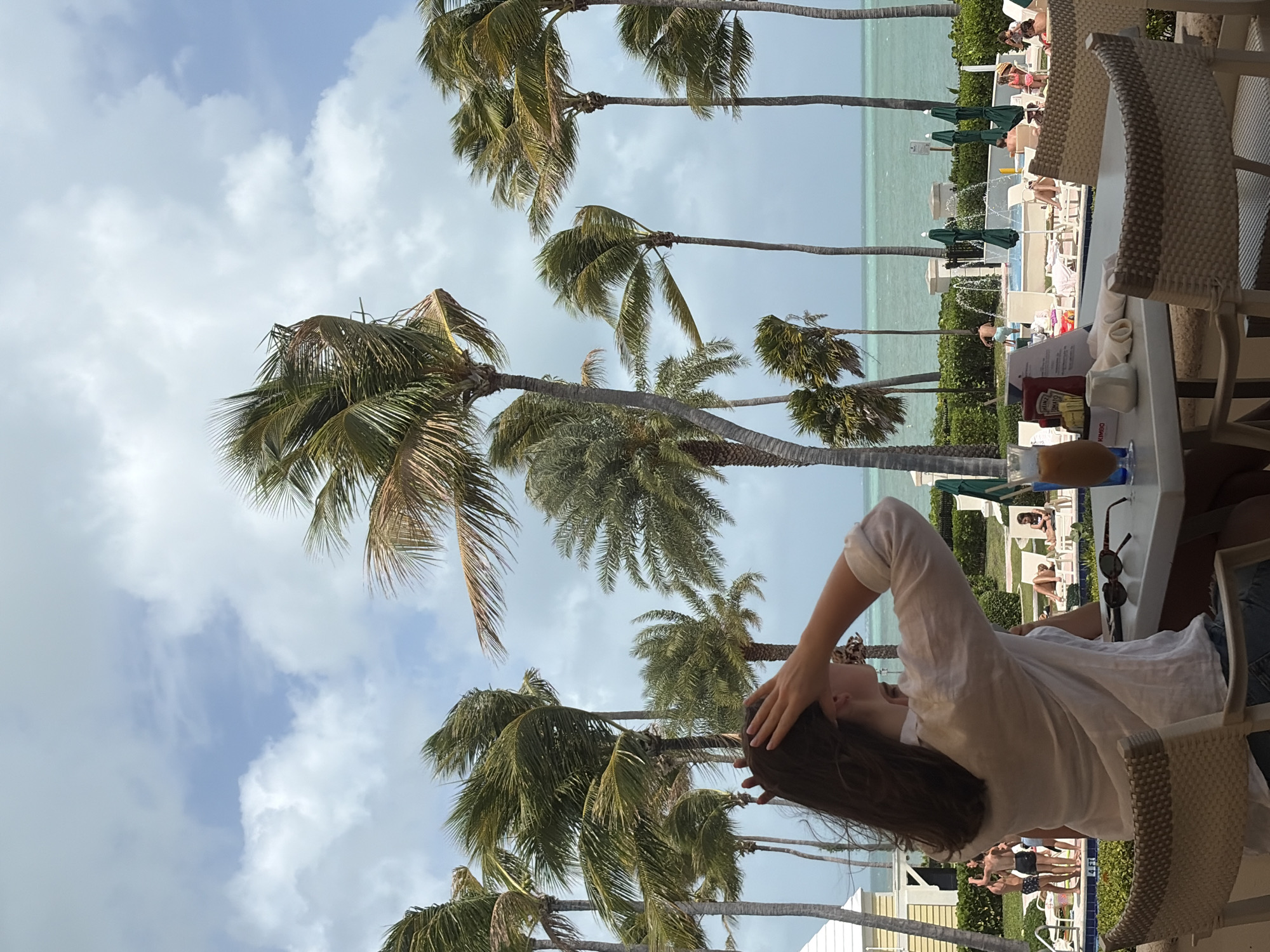 A horizontal, eye-level shot captures a scene at the beach in Key Biscayne, Florida. Palm trees line the shore, with the ocean visible in the background, alongside sunbathers in lounge chairs. A woman is visible, seated at a table, with her hand on her head. The table also contains items like a menu and sunglasses.