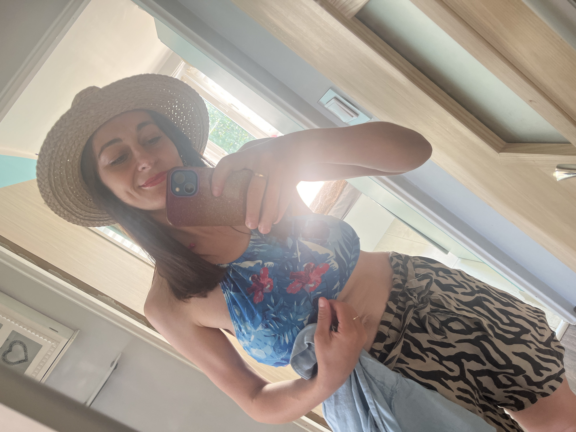 A woman is taking a mirror selfie in what appears to be a doorway. She is wearing a straw hat, a blue and floral print crop top, and a patterned skirt with a zebra-like print. She is holding a phone in her right hand, taking the picture, and her left hand is holding a piece of light blue fabric. The lighting suggests it is daytime, possibly morning or afternoon, with sunlight streaming in. The background shows a wooden doorframe and a glimpse of what could be a room interior with some decorative items. The woman has red lipstick on and appears to be smiling.