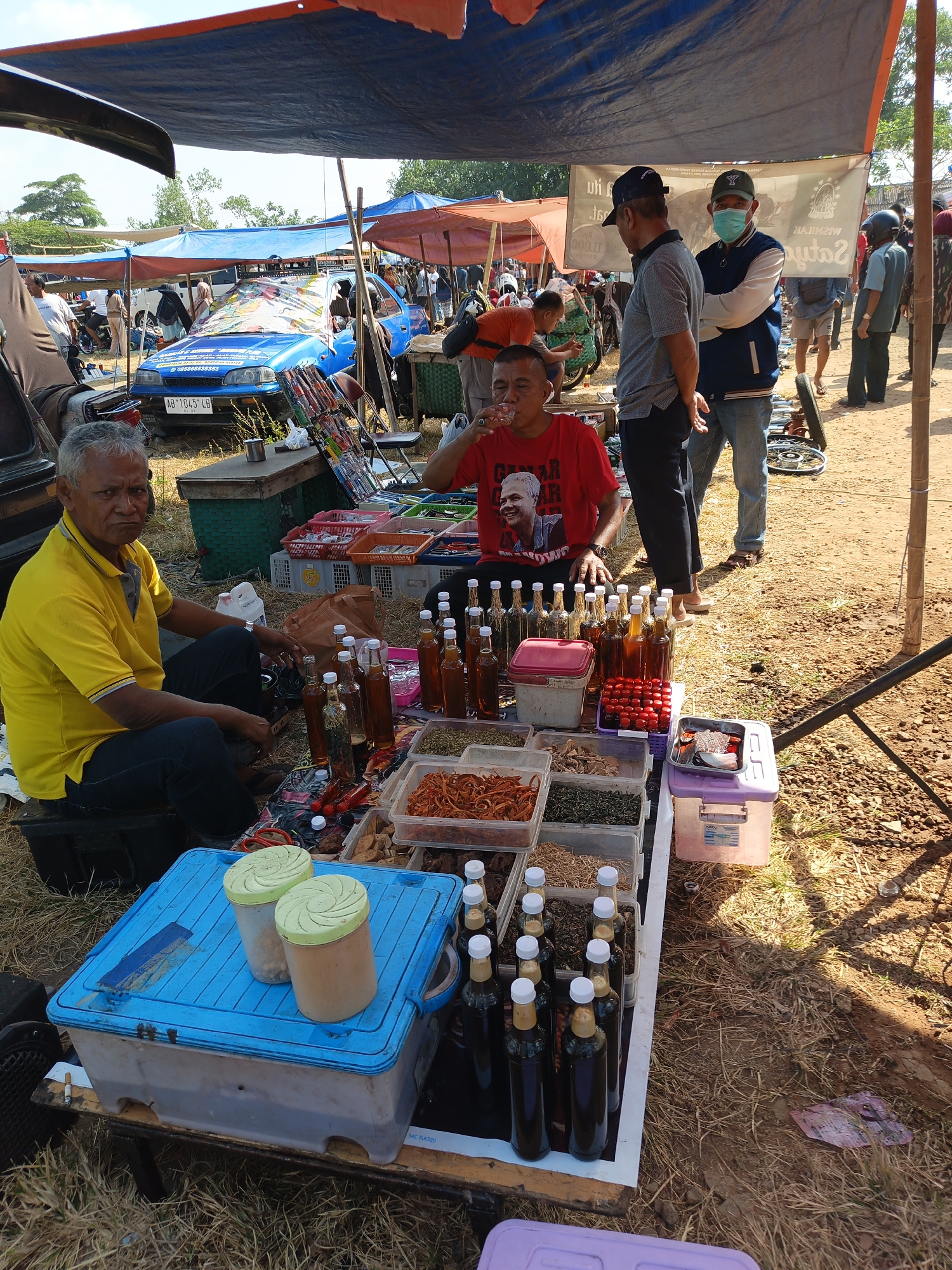 This is a forensic, matter-of-fact description of a scene in Jatinom, Indonesia.
The image captures a lively outdoor market scene, likely a roadside or open-field market, under a makeshift canopy. A man wearing a bright yellow polo shirt and dark pants sits on a black plastic stool, facing left and looking directly at the camera. He appears to be a vendor, with his hands resting on a table filled with various goods.
The table is laden with an assortment of items, including numerous glass bottles filled with dark liquid, possibly traditional medicine or beverages, and plastic containers holding dried herbs, spices, or food items. Two large, light green lidded containers are placed on top of a blue plastic storage box in the foreground. A red plastic container with small red items, possibly berries or candies, is also visible.
In the background, other individuals are present, some browsing or walking through the market. A blue car with a colorful tarp draped over its windshield is parked behind the stall. Other market stalls and canopies are visible, indicating a bustling commercial environment. The ground is covered with dry grass and soil, typical of an outdoor setting in a rural or semi-rural area. The overall atmosphere suggests a daytime market, possibly in the midday sun given the strong shadows. The specific location is Jatinom, Indonesia.