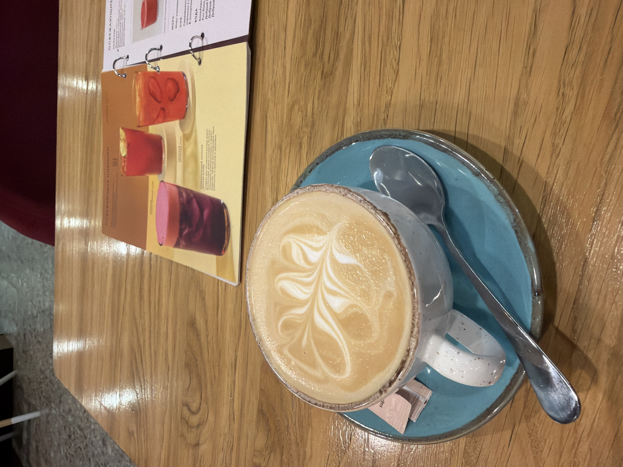 A close-up, high-angle shot captures a ceramic mug of latte art atop a teal-colored saucer, resting on a polished wooden table. The latte art features a symmetrical, swirling design in white foam against the creamy coffee. A metal spoon lies across the saucer, partially obscured by the mug's handle. To the left of the mug, a few small, pink sugar packets are visible. In the background, a menu is laid open, displaying images of colorful drinks. The lighting is warm and diffused, creating reflections on the table surface. The scene appears to be set in a cafe or restaurant.