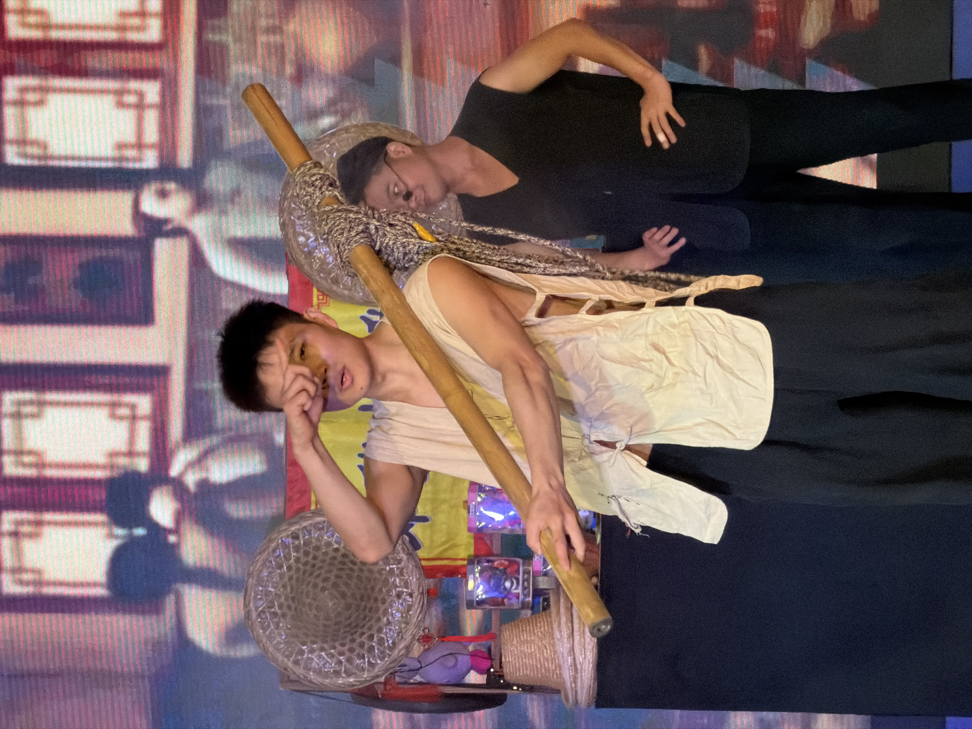 The image shows two men on a stage, likely during a performance. The man in the foreground is holding a long wooden pole across his shoulders, with his right hand touching his forehead. He is wearing a loose-fitting, light-colored sleeveless tunic over dark pants. To his right, a second man stands with his hands on his hips. He is wearing a black vest over a black shirt and dark pants, and appears to be wearing glasses. Behind them, a large screen displays an image of people in traditional clothing, possibly an artistic backdrop. There are also props such as straw hats and decorative items on display in front of the screen. The lighting suggests an indoor venue. The overall scene depicts a theatrical or cultural performance.