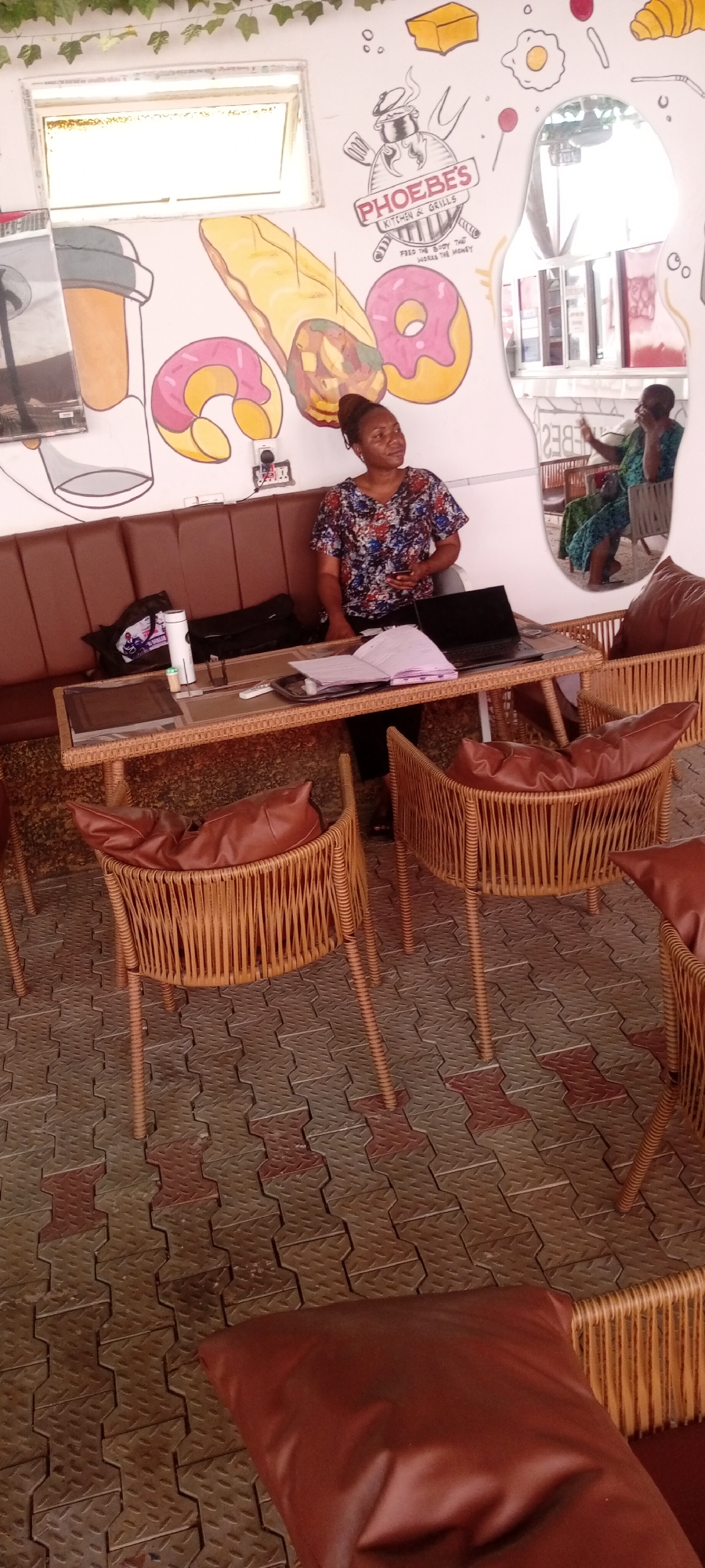 The image shows the interior of a café named "Phoebe's Kitchen & Grills" located in Sabon Gari East, Nigeria. A woman in a floral shirt is standing behind a wicker table with a laptop, a book, and other items. The table is surrounded by wicker chairs with brown leather cushions. Behind her is a brown leather bench and a wall with a mural of food items such as doughnuts and a burrito. A mirror reflects a seated woman. The floor is paved with interlocking concrete blocks.