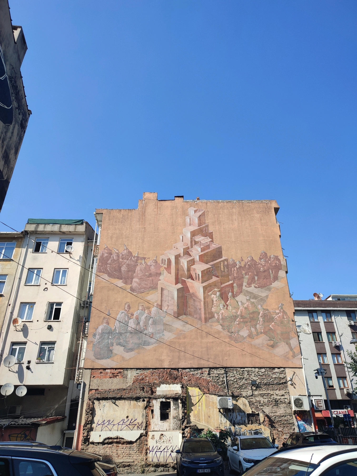 The image shows a mural on the side of a building in Kadıköy, Turkey. The mural depicts a scene with several figures dressed in historical attire, possibly representing monks or scholars, gathered around a complex architectural structure made of stacked cubes. The building itself appears to be aged, with some areas of exposed brickwork and graffiti near the base. Adjacent to the muraled building is a lighter-colored, multi-story apartment building with numerous windows and satellite dishes. In the foreground, several cars are parked on the street, including a dark SUV, a white sedan, and a white car with its roof visible. The sky is clear and blue, suggesting it is daytime.