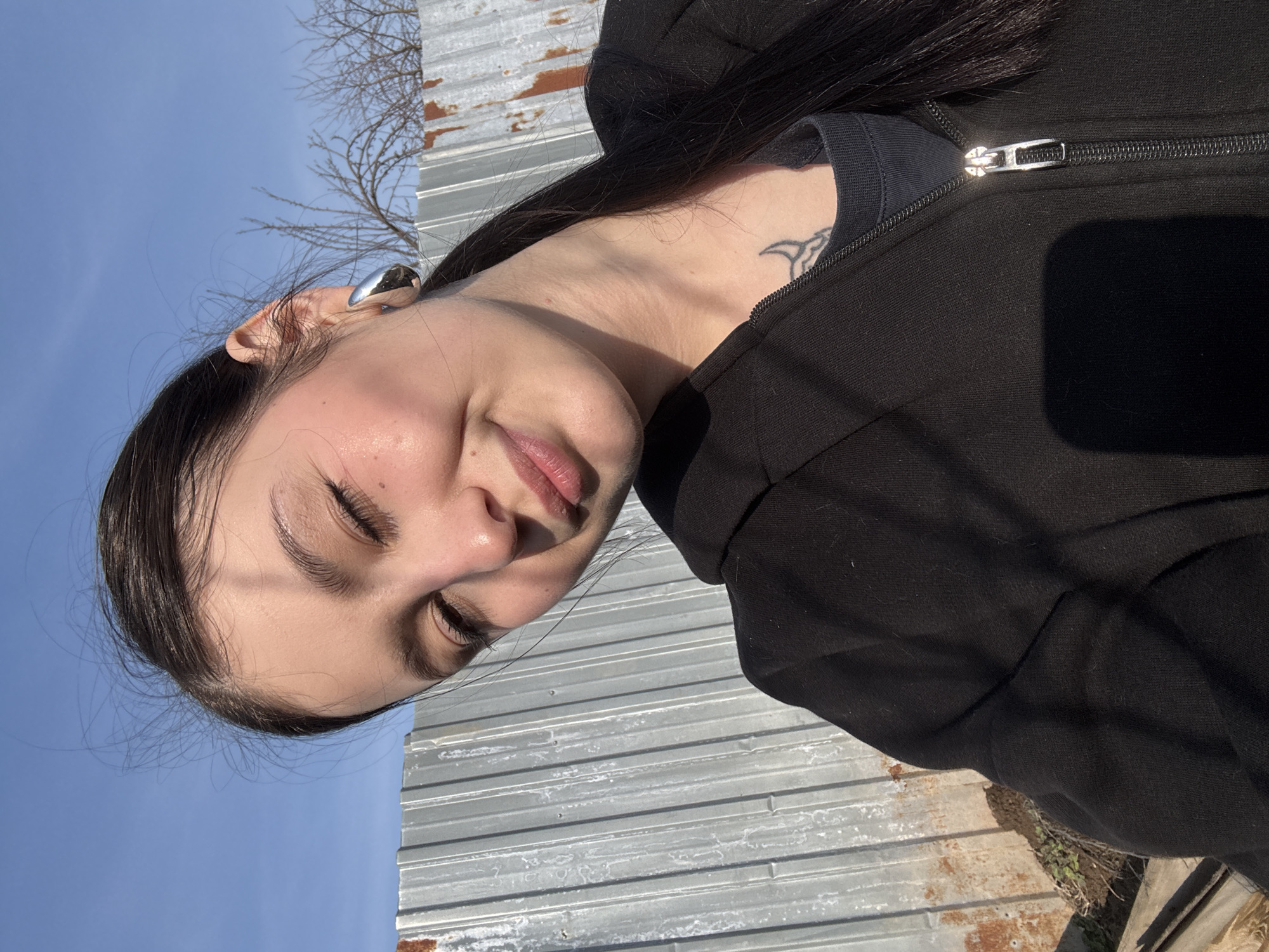 A rotated, close-up portrait depicts a light-skinned female wearing a black zip-up hoodie over a dark shirt. She has dark hair, with a silver ear adornment. A tribal-style tattoo is visible on her neck. The background features corrugated metal, bare branches, and a clear blue sky. The location is near Фоки, Russia.