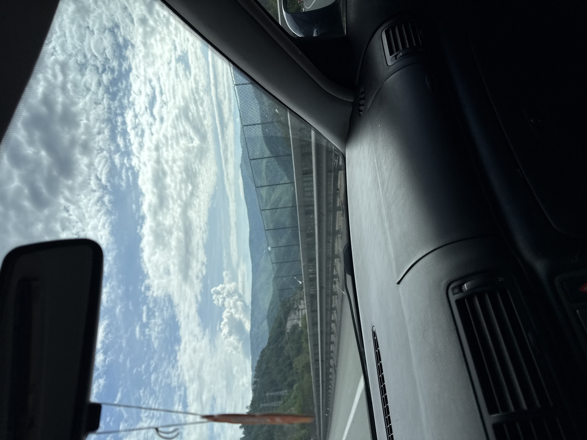 The image, oriented vertically as if rotated onto its side, captures a scenic view from within a vehicle, looking out through the front windshield.
Outside the car, a highway or multi-lane road with white lane markings is visible, running parallel to a tall, dark chain-link fence. Beyond the fence, a landscape of verdant mountains covered in dense forests stretches across the mid-ground and background. The sky above is a bright blue, filled with numerous fluffy, white cumulus clouds, suggesting a clear and sunny day.
Inside the vehicle, the dark dashboard occupies the right side of the frame, featuring textured surfaces and visible air conditioning vents. A small portion of the rearview mirror is visible on the far left, reflecting parts of the sky. A slender, reddish-brown object, possibly an air freshener or decorative item, hangs down near the top-left corner of the view.
The scene depicts a journey or travel by car through a mountainous region, consistent with the location context of Gorno Kamartsi, Bulgaria. The activity is passive observation from a moving vehicle. The time of day is clearly daytime, characterized by bright sunlight and visible clouds. No discernible text is present in the image.