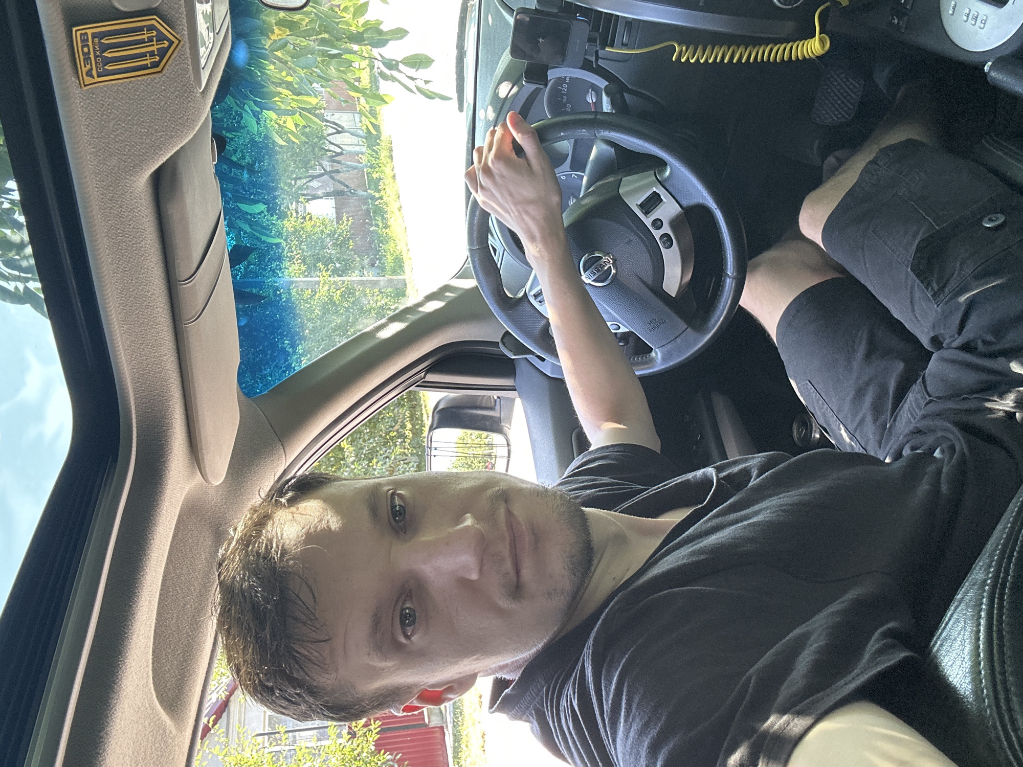A man is sitting in the driver's seat of a car. The car's sunroof is open, allowing natural light to illuminate the interior. A decal featuring three golden trident symbols on a blue background with the text "АЗОВ ССО КИЇВ" is affixed to the car's interior ceiling. The man is wearing a black t-shirt and black shorts. He is looking directly at the camera. His right hand is resting on the steering wheel. The steering wheel has the Nissan logo in the center. The car's dashboard and center console are visible. Outside the car, trees and a building are visible, suggesting an outdoor setting. The presence of the decal with "АЗОВ ССО КИЇВ" indicates a possible connection to Ukraine. The context provided indicates the location is Shatava, Ukraine.