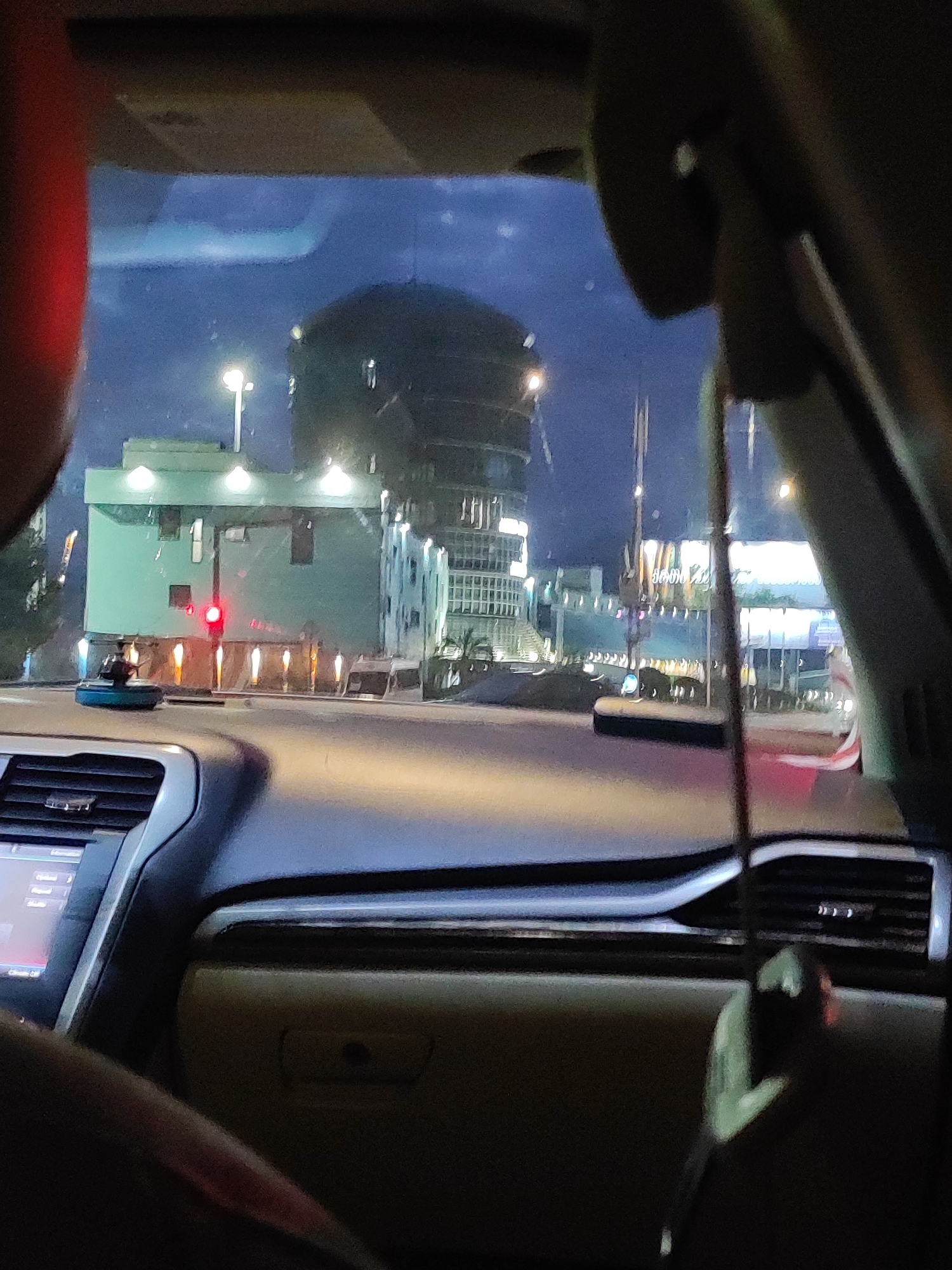 The image captures a view from inside a car looking out at a cityscape at dusk or night, with a focus on a rounded, multi-story building in the background and a lower, green building in the mid-ground. The green building features a red traffic light and several evenly spaced vertical lights along its facade. The rounded building appears to be illuminated from within or by external lights, showcasing a textured exterior. In the foreground, the dashboard of the car is visible, with a navigation screen on the left and a light-colored dashboard shelf on the right. The overall scene suggests an urban environment, possibly in Batumi, Georgia, given the architectural style and lighting. The perspective implies the car is stopped at a traffic light. There are no discernible individuals in the scene, but the presence of the car and the urban setting indicate human activity.