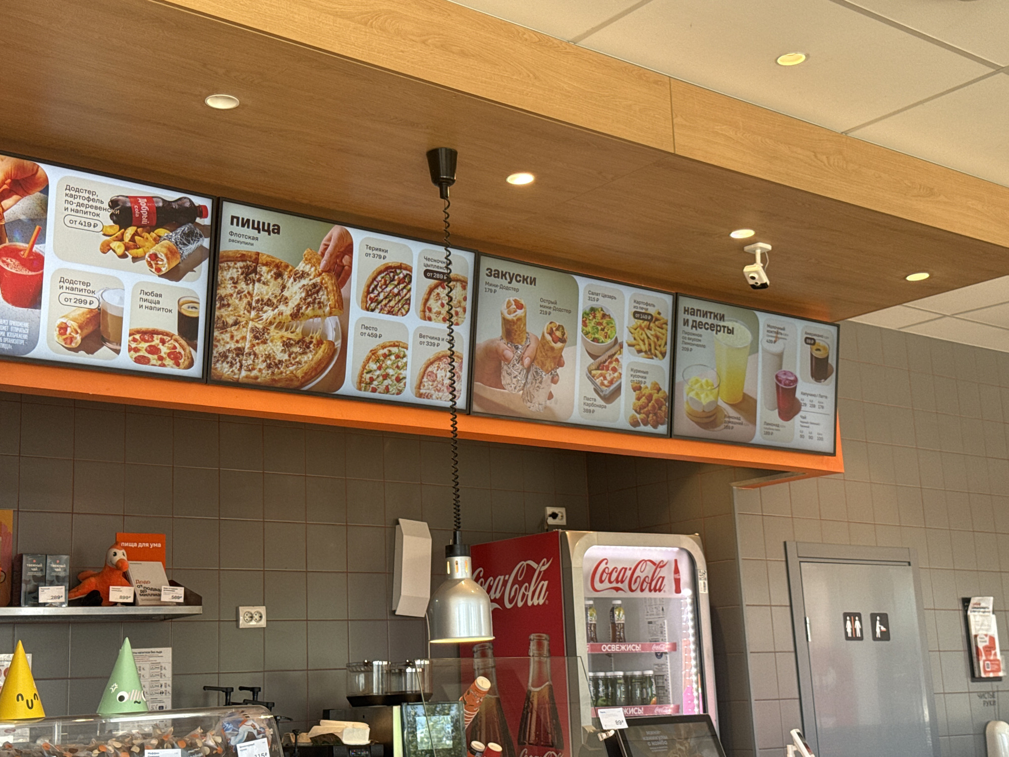 The image shows the interior of a fast-food restaurant, likely a pizza place, with a menu board displayed prominently. The menu board features various food and drink options with prices in Russian rubles.
**Subjects:** The primary subjects are the food items advertised on the menu board, including pizzas, dodsters (a type of wrap or sandwich), fries, chicken pieces, salads, and drinks. There are no people clearly visible in the image, though there are pictograms of people on a gray door in the background. Several objects are visible: a Coca-Cola refrigerator, a suspended lamp, decorative cones on a counter, and stuffed toys on a shelf.
**Scene and Setting:** The scene is set within a bright, modern fast-food establishment. The walls are tiled in gray, and the ceiling is a light wood paneling with recessed lighting. The overall atmosphere appears clean and organized, typical of a commercial food service environment. The Coca-Cola branding is visible on the refrigerator and the menu board, suggesting a partnership or the sale of Coca-Cola products.
**Activity or Event:** The image captures a moment of retail display, showcasing the offerings of the restaurant. The menu board is active, implying that customers are either viewing it or ordering from it.
**Notable Details:**
* **Time of Day:** The bright lighting suggests it is daytime.
* **Location Cues:** The Russian text on the menu board ("пицца," "напитки," prices in "Р") and the location context "Golitsyno, Russia" confirm the locale.
* **Visible Text:** The menu board displays item names like "пицца," "Додстер," "закуски," "напитки и десерты," along with prices. The Coca-Cola logo is also clearly visible.
The image provides a glimpse into the visual presentation and offerings of a fast-food restaurant in Russia.