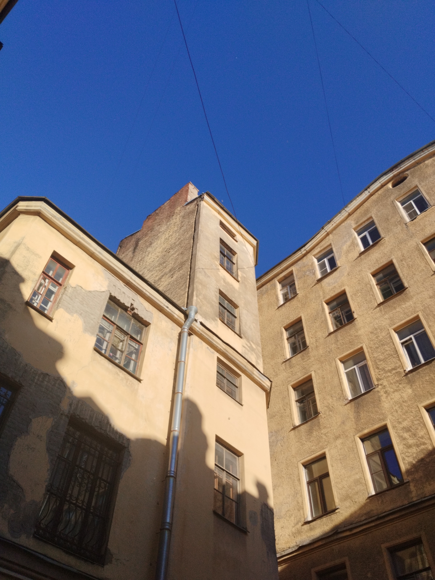 The media file shows a view looking up at the corner of two buildings in Saint Petersburg, Russia, against a bright blue sky. The buildings are old, with yellow-beige stucco exteriors that show signs of wear and tear, including peeling paint and some areas of exposed brick. The architecture suggests a courtyard setting, common in historic Saint Petersburg.
The left building is partially in shadow, revealing architectural details like ornate window frames and a barred window on a lower floor. A drainpipe runs vertically down the side of this building. The right building is fully illuminated by sunlight, showcasing a pattern of numerous windows. These windows are of various sizes and styles, some with white frames and others with darker frames, and some appear to have reflective glass. The angle of the buildings and the shadows suggest it is midday.
Thin wires, likely power lines or telephone lines, stretch across the clear blue sky, a common sight in urban environments. The overall impression is one of an older, well-established urban environment, possibly a residential or administrative area. There are no people or specific activities depicted, making it a still-life shot of the urban architecture. The lighting indicates a clear, sunny day.