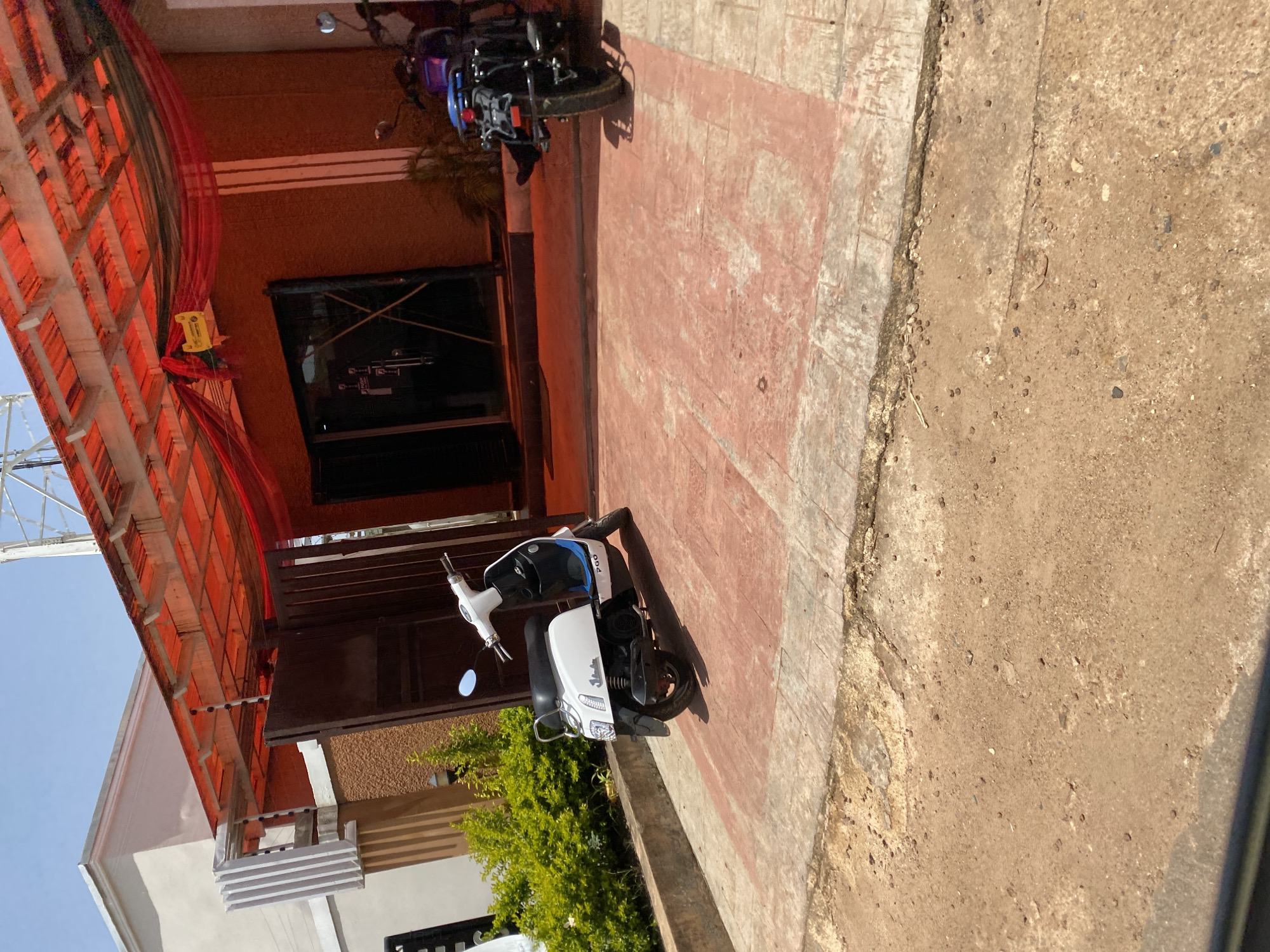 This media file shows an outdoor scene in Gura Top, Nigeria. The image is oriented vertically, showing a building entrance with a red awning overhead. In front of the building, there are two motorbikes parked on a red tiled pathway. The first motorbike, closer to the foreground, is white with blue and black accents. The second motorbike, further back, is black with purple accents.
The building itself appears to have an orange-brown facade with a dark entranceway. To the left of the building, a tall white structure, possibly a radio tower, is partially visible against a bright blue sky, suggesting a clear day. Green foliage is visible near the base of the building, next to the white motorbike.
The ground is a mix of red tiles and rough, sandy concrete. The lighting suggests it is daytime, with sunlight casting shadows from the motorbikes and awning. There is no visible text in the image. The scene depicts a quiet moment, possibly in front of a residence or a small business.