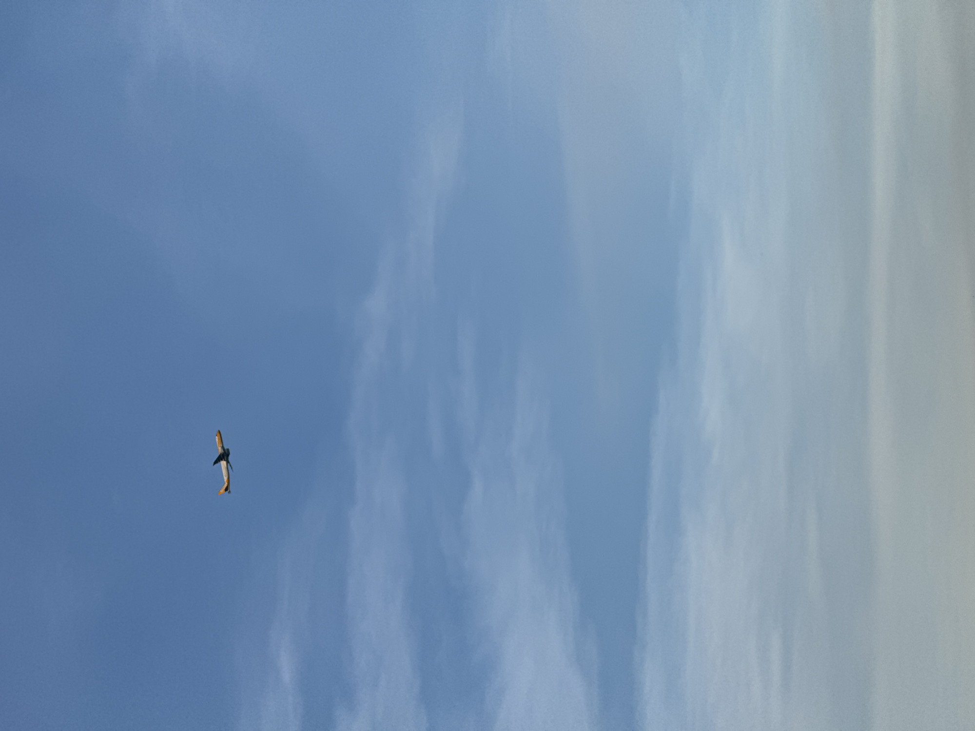 An airplane with an orange and white livery is flying in a clear blue sky with wispy clouds. The airplane is positioned in the upper left quadrant of the image, ascending. The horizon is not visible.