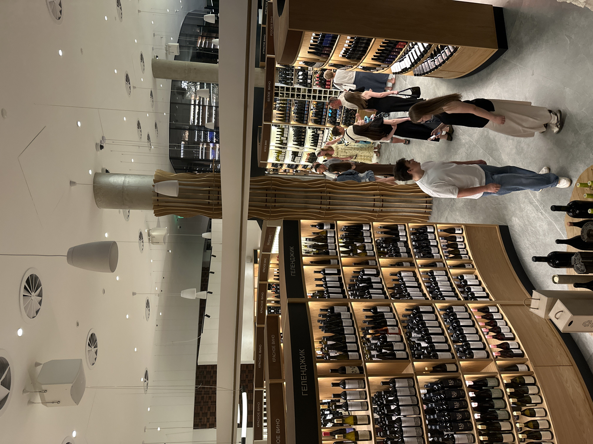 The image depicts the interior of a modern wine store, identified as being located in Gelendzhik, Russia, based on signage visible in the photograph. The store features extensive shelving units filled with bottles of wine, organized by type and origin, indicated by signs such as "КРАСНОЕ ВИНО" (Red Wine), "БЕЛОЕ ВИНО" (White Wine), and "ГЕЛЕНДЖИК".
Several individuals are present in the store, browsing the selections. In the foreground, a young man in a white t-shirt and blue jeans is talking to a young woman wearing a black top and a long, beige skirt. Other shoppers are visible throughout the aisles, some examining bottles, others walking or standing in groups. The store's architecture is contemporary, with high ceilings, integrated lighting, and decorative wooden elements. Large windows in the background offer a glimpse of the exterior or additional sections of the store, showcasing more wine displays. The overall atmosphere appears to be that of a well-stocked and upscale retail establishment.