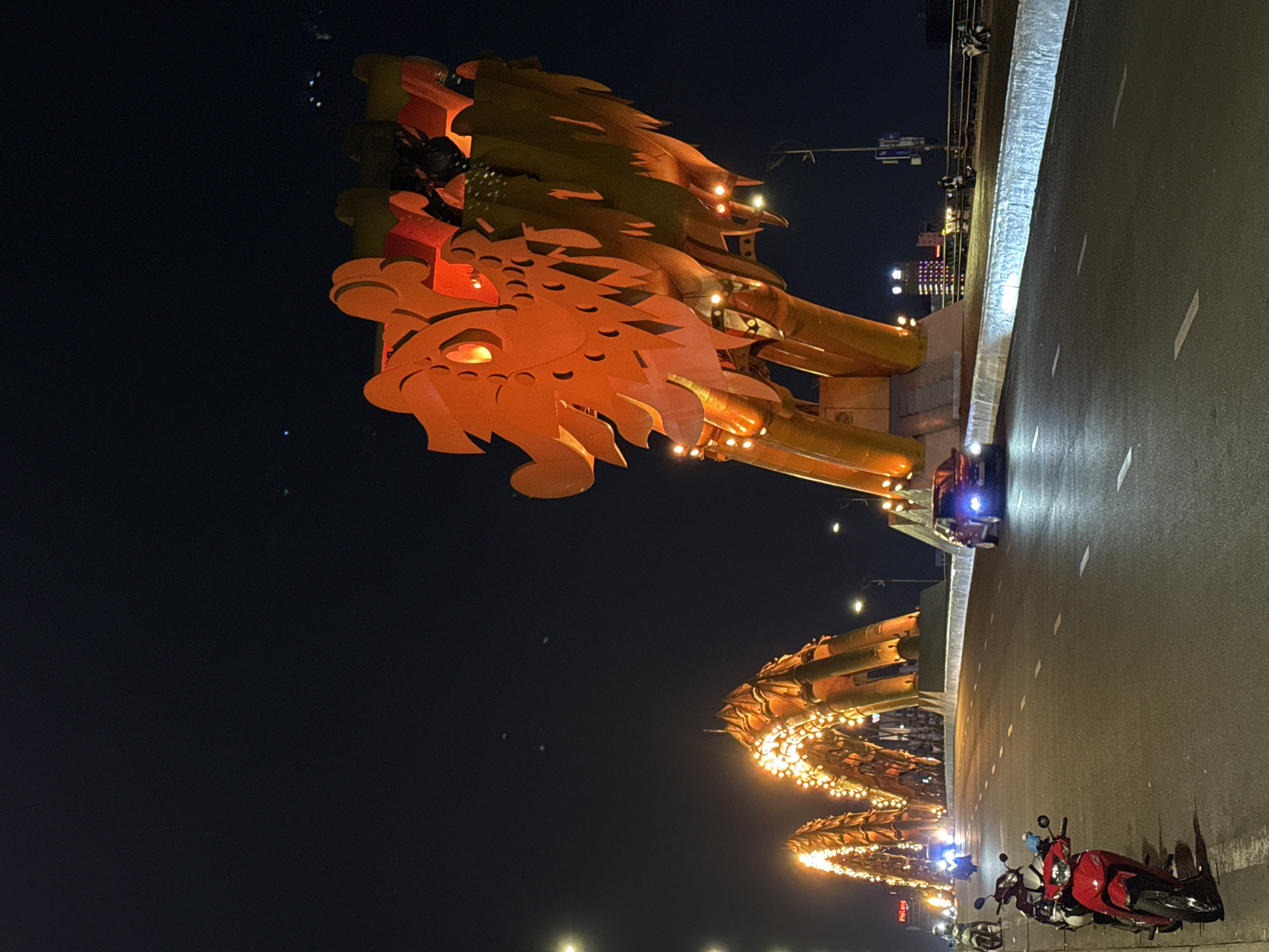 The image displays a vibrant night scene of the Dragon Bridge in Da Nang, Vietnam. The main subject is the intricately designed head of a large, golden-orange dragon sculpture, illuminated by numerous warm lights, positioned on the left side of the frame. Its scales and features are clearly visible against the dark sky.
Stretching from the dragon's head across the frame, segments of the dragon's body are visible, forming the bridge's structure and also brightly lit by countless small, warm-toned lights, creating a shimmering effect. On the right side of the image, the paved road surface of the bridge is visible, with distinct white lane markings. A red scooter or motorbike is parked on the side of the road in the lower right corner. Further down the bridge, another dark-colored vehicle and a dimly visible person can be seen.
The time of day is clearly night, indicated by the dark sky and the extensive artificial illumination of the bridge and distant city lights. The weather appears clear. The prominent dragon structure immediately identifies the location as the famous Dragon Bridge in Da Nang. On a sign mounted on the bridge structure, beneath the illuminated dragon's body, some red text is visible, partially reading "Phuong H...". The overall atmosphere is one of urban nightlife and architectural spectacle.