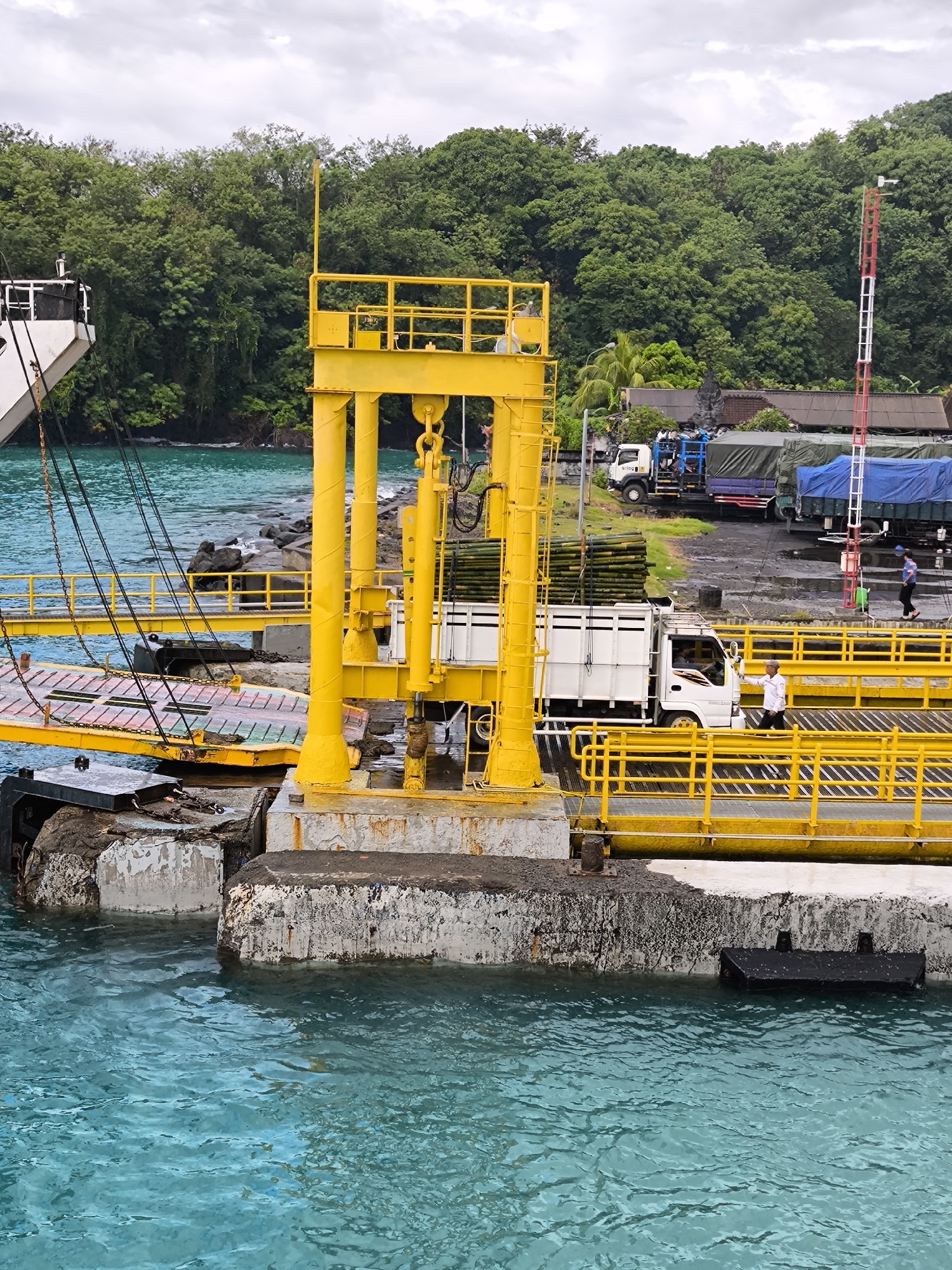 The image depicts a ferry terminal in an unspecified location. In the foreground, a yellow metal framework supports a ramp extending towards a docked vessel. The water appears turquoise, with concrete structures partially submerged. A white truck loaded with green lumber is driving across the yellow ramp. A man dressed in white directs the truck. In the background, other trucks are parked on a tarmac area, partially obscured by lush green trees. A red and white striped tower is visible. A few individuals are walking on the tarmac. The sky is overcast, suggesting a cloudy day.