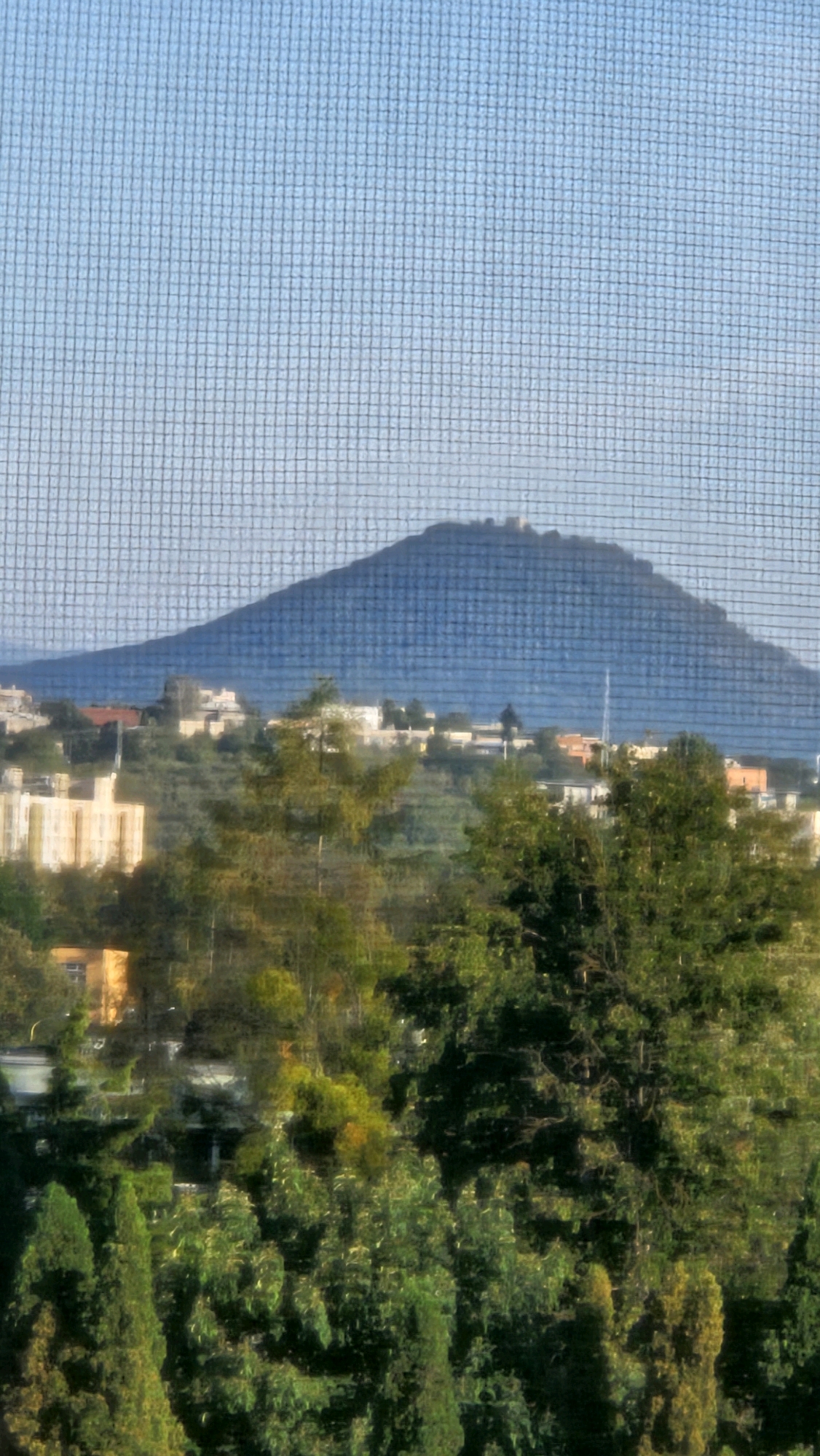 The image, taken through a mesh screen, depicts a landscape dominated by a large, bluish-grey mountain in the background. The mountain has a flat top with some discernible structures, possibly ancient ruins or fortifications. In the midground, a town or village is nestled on the slopes leading up to the mountain. Buildings with light-colored facades and some orange roofs are visible. Several trees, varying in size and shade of green, are scattered throughout the midground and foreground, some appearing denser than others. The sky is a light blue with a textured appearance, consistent with being viewed through a screen. The overall impression is of a sunlit, somewhat hazy view of a rural or semi-urban area with a prominent natural landmark. This scene is identified as the Zemplín Castle (Zemplínsky hrad) and the surrounding area near the city of Prešov, Slovakia.