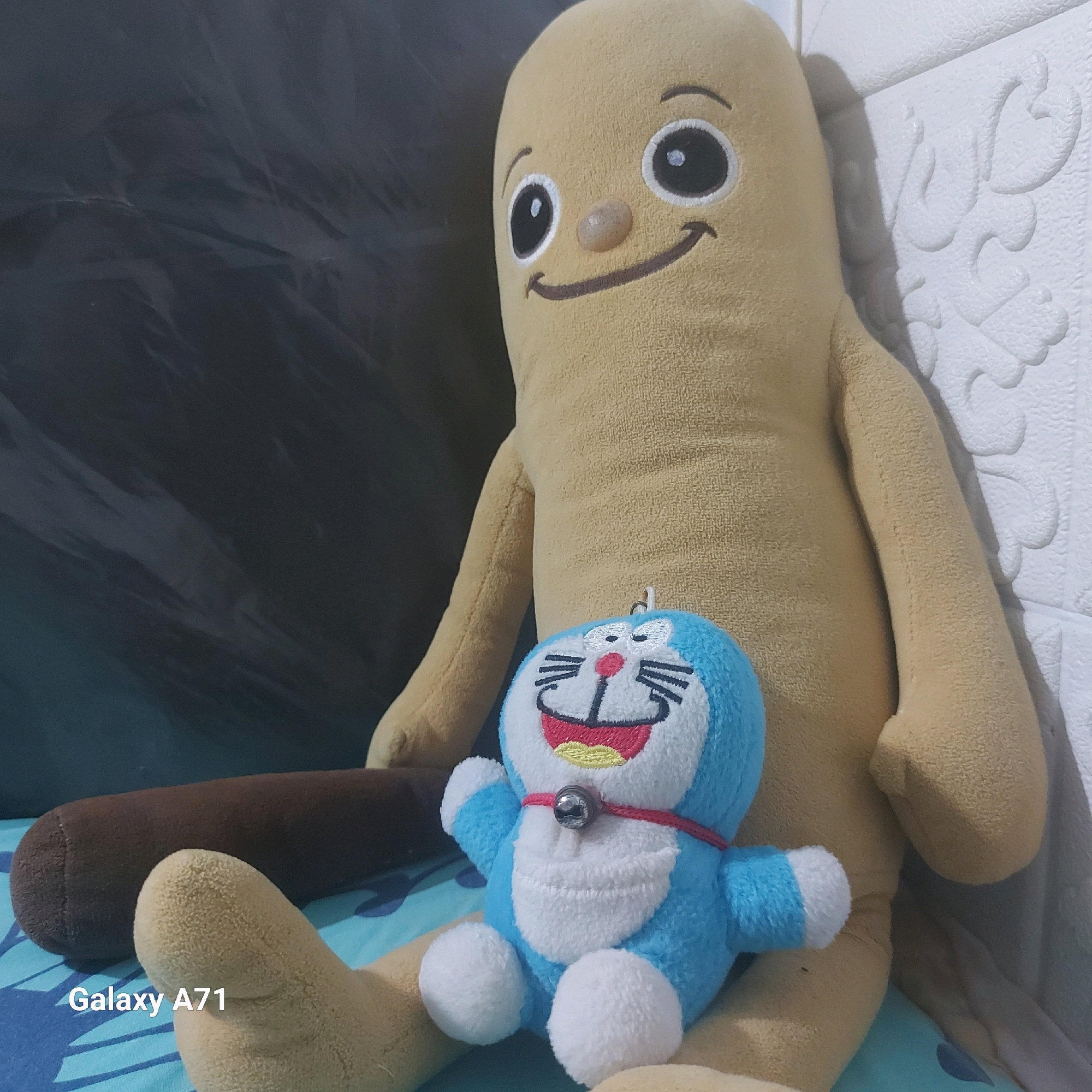 The image shows two plush toys, one large and yellow with a round face and simple smile, and a smaller blue and white plush toy resembling Doraemon, with a bell around its neck, positioned in front of the larger toy. A brown cylindrical object, possibly another toy, lies horizontally behind the Doraemon plush. The toys are placed on a surface with a blue and white patterned fabric. In the background, a white textured wall is visible on the right, with a dark, possibly fabric, background on the left. The bottom left corner of the image shows the text "Galaxy A71", indicating the device used for capturing the photograph. The scene appears to be indoors, likely in a residential setting. There is no specific information in the image to determine the location within the city of Surabaya, Indonesia.