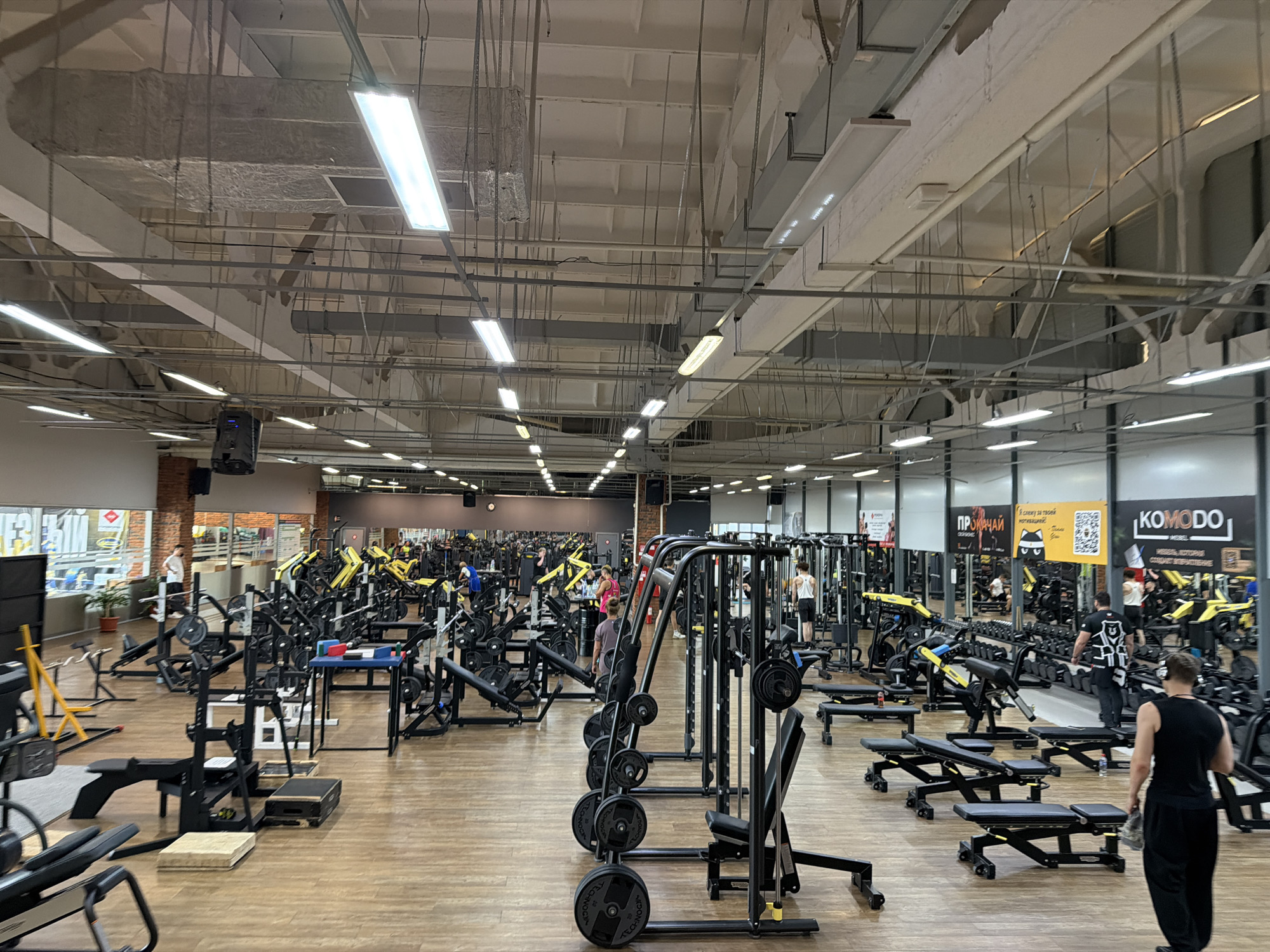 This is an interior shot of a large, modern gym with light wooden floors and a high ceiling. Fluorescent lights are visible along the ceiling, along with exposed ventilation and wiring. The gym is filled with various weightlifting machines and benches, many of which are black with yellow accents. Several people are present, scattered throughout the gym, some are working out on machines, while others are walking or observing. In the background, there are windows showing glimpses of an urban or industrial setting. Signs in Russian are visible on the walls, including one that reads "KOMODO." The overall impression is a well-equipped fitness facility. The location is identified as Chaikovsky, Russia.