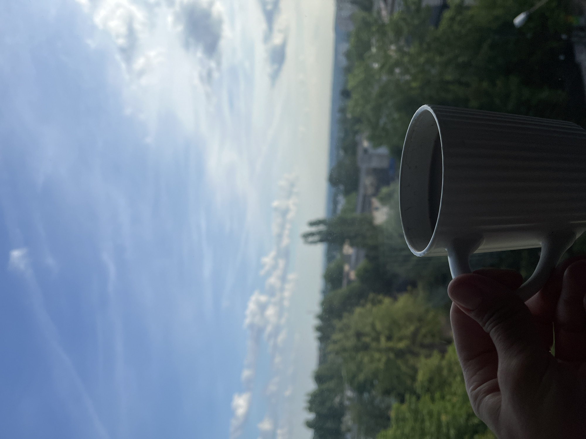 A close-up shot shows a person's hand holding a white mug filled with a dark liquid, likely coffee, against a blurry backdrop of a city skyline and a bright, cloudy sky. The mug has a ribbed texture. The hand is positioned in the lower left quadrant of the frame, obscuring the view of the lower portion of the mug. The background depicts a dense canopy of green trees interspersed with buildings, suggesting an urban or suburban environment. The sky is a pale blue with scattered white clouds, and the overall lighting suggests it is daytime. The specific location within the city of Unknown, Unknown cannot be determined from the visual information provided.
