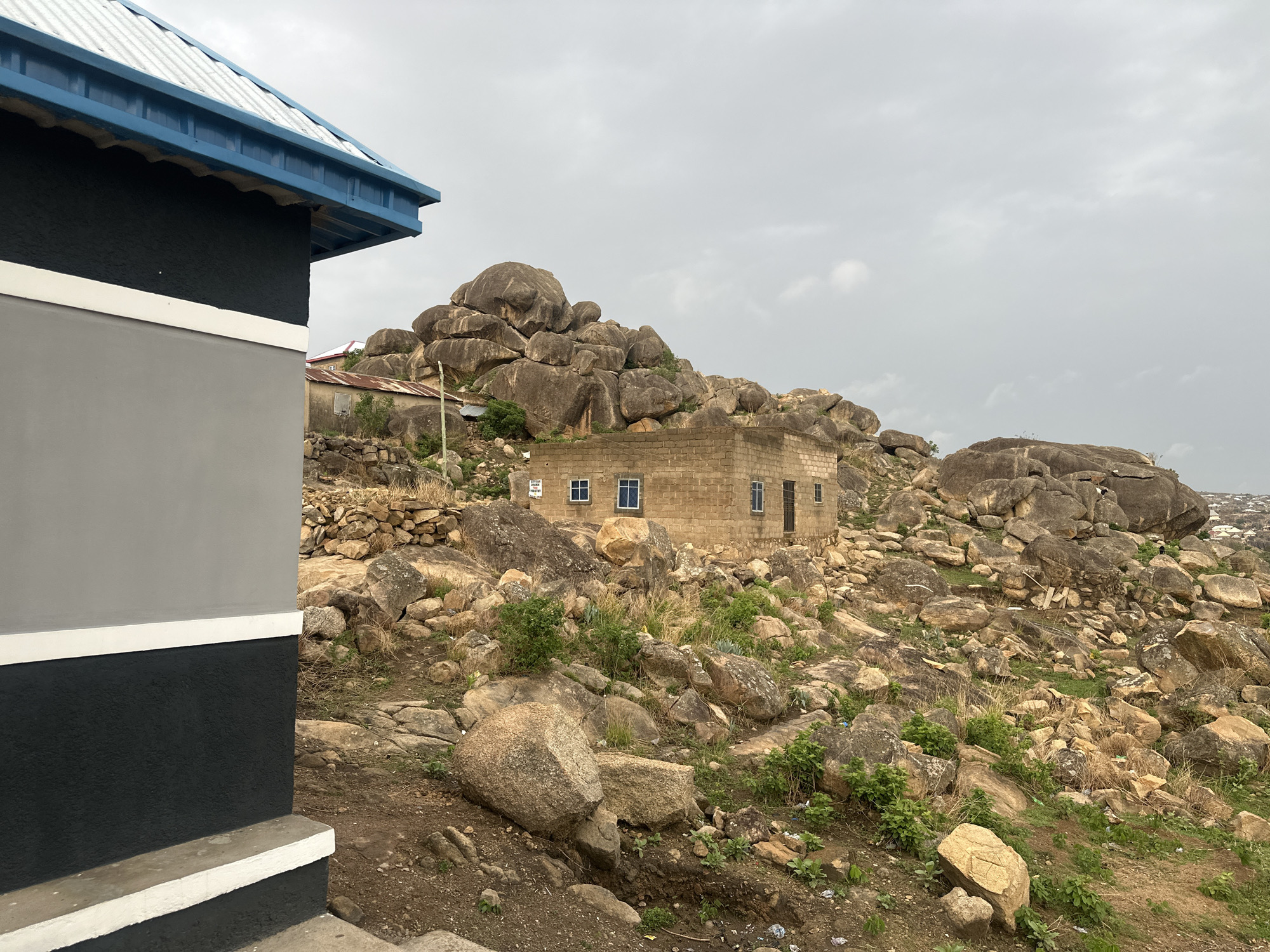 The image shows a hillside scene in Jos, Nigeria, with a partially constructed building in the foreground to the left. The building has a gray and black exterior with white trim and a blue roof edge. The hillside is rocky, covered in various sizes of tan and gray boulders. Vegetation, consisting of sparse grass and small bushes, is scattered among the rocks. In the middle ground, a rectangular brick building with a corrugated metal roof is nestled among the boulders. A cluster of large, rounded boulders forms a backdrop to the buildings. The sky is overcast and gray. In the distance, the urban landscape of Jos is faintly visible on the right side of the frame.