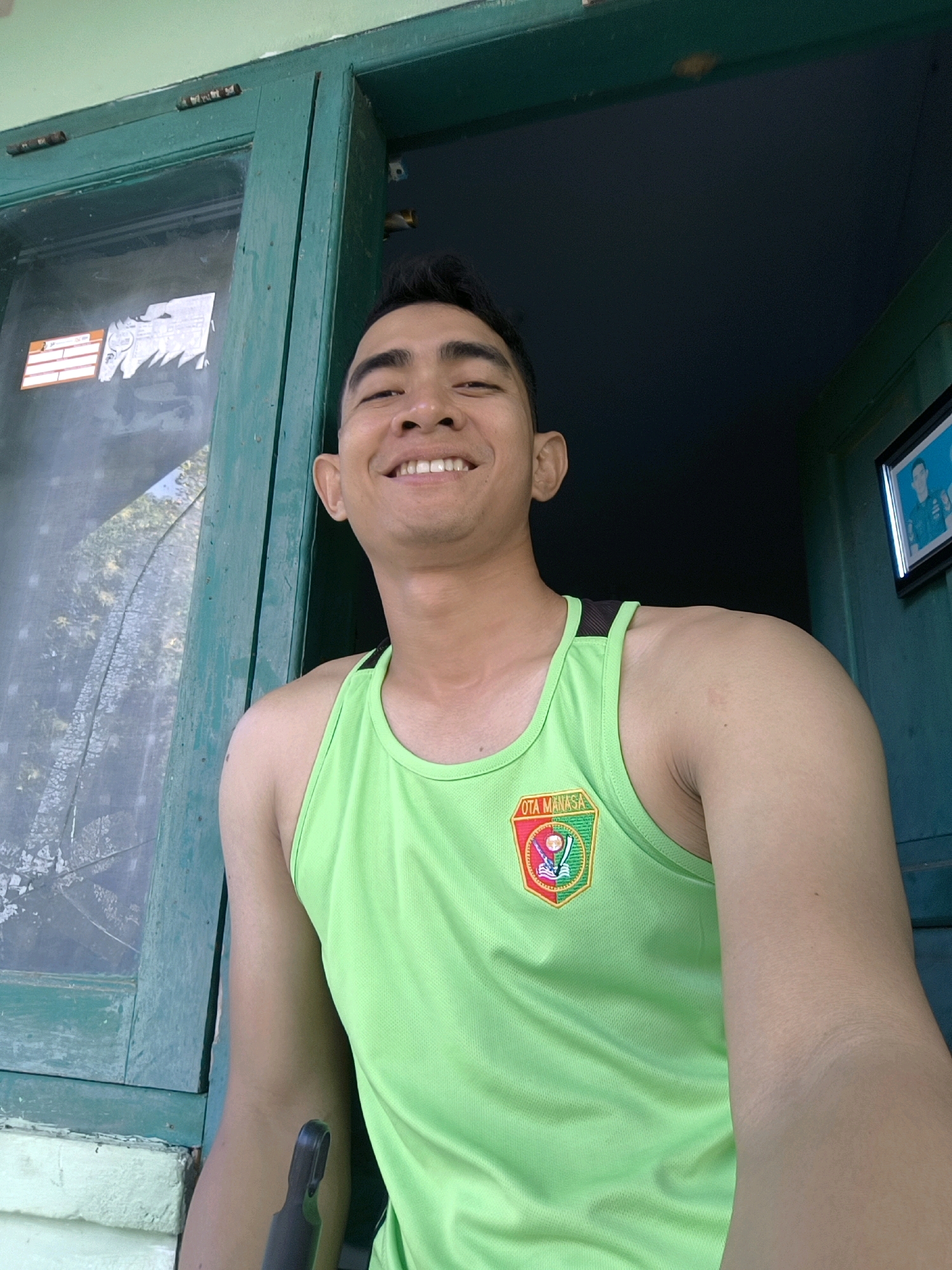 An outdoor selfie features a young man in a bright green tank top. The man has dark hair styled upwards and a visible smile. He is positioned in front of a weathered green wooden door frame. To his left, a glass pane shows a distorted reflection of trees and sky. A small, colorful crest with the text "OTA MANASA" is visible on the left side of his tank top. In the background, to the right of the man, a framed picture hangs on a dark blue wall. The scene appears to be in a residential or public area, possibly in Indonesia, given the style of the building and the text on the crest.