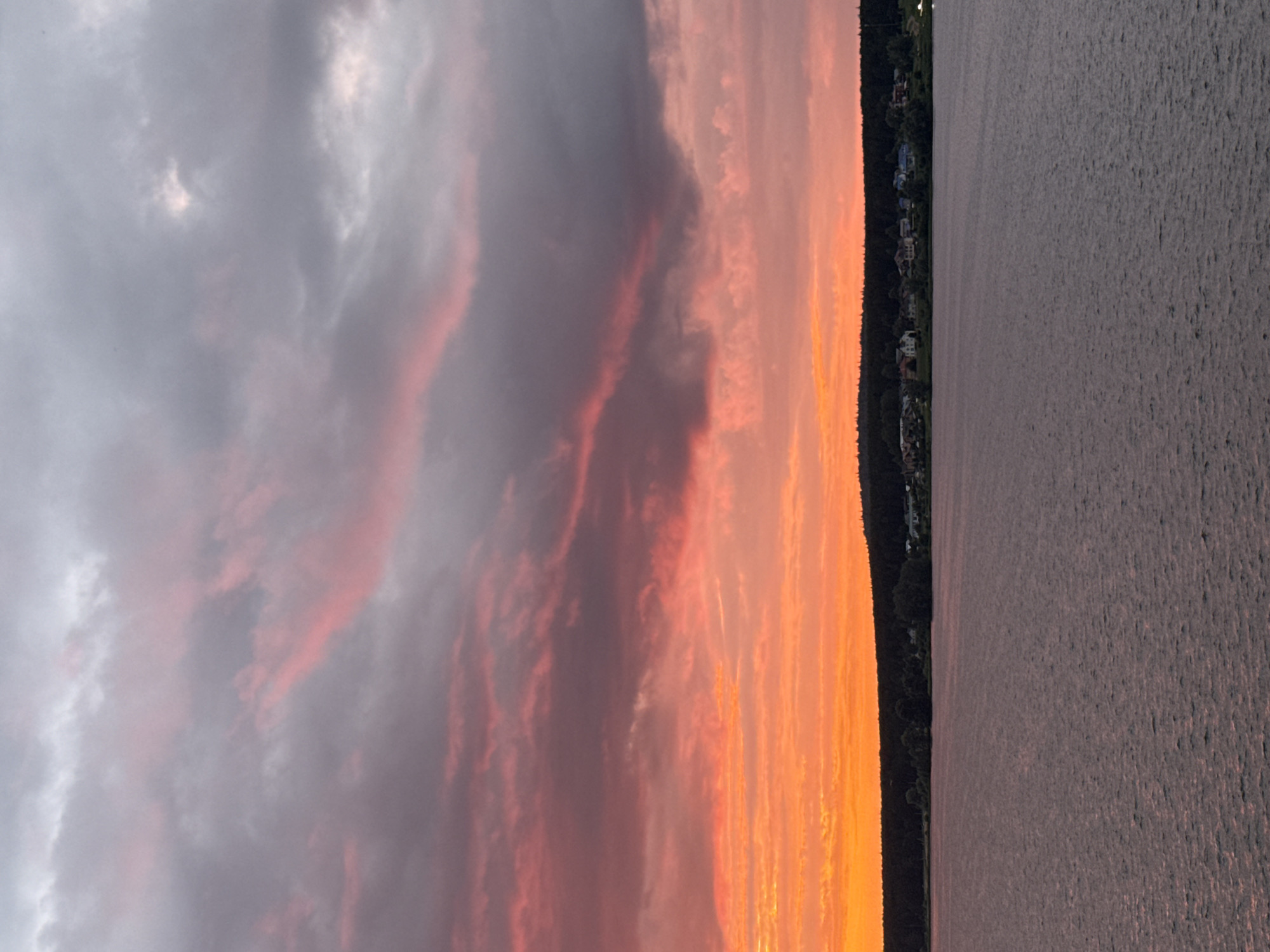 The image depicts a sunset over a body of water, likely a lake or river, in Votkinsk, Russia. The sky is filled with dramatic clouds, transitioning from a bright orange and pink hue near the horizon to darker grays and purples higher up. A dark line of trees forms the shoreline, with a scattering of houses visible nestled amongst them. The water in the foreground is calm, with a rippled surface reflecting the colors of the sky. No people or specific actions are visible in the scene, which appears to be a natural landscape at dusk.