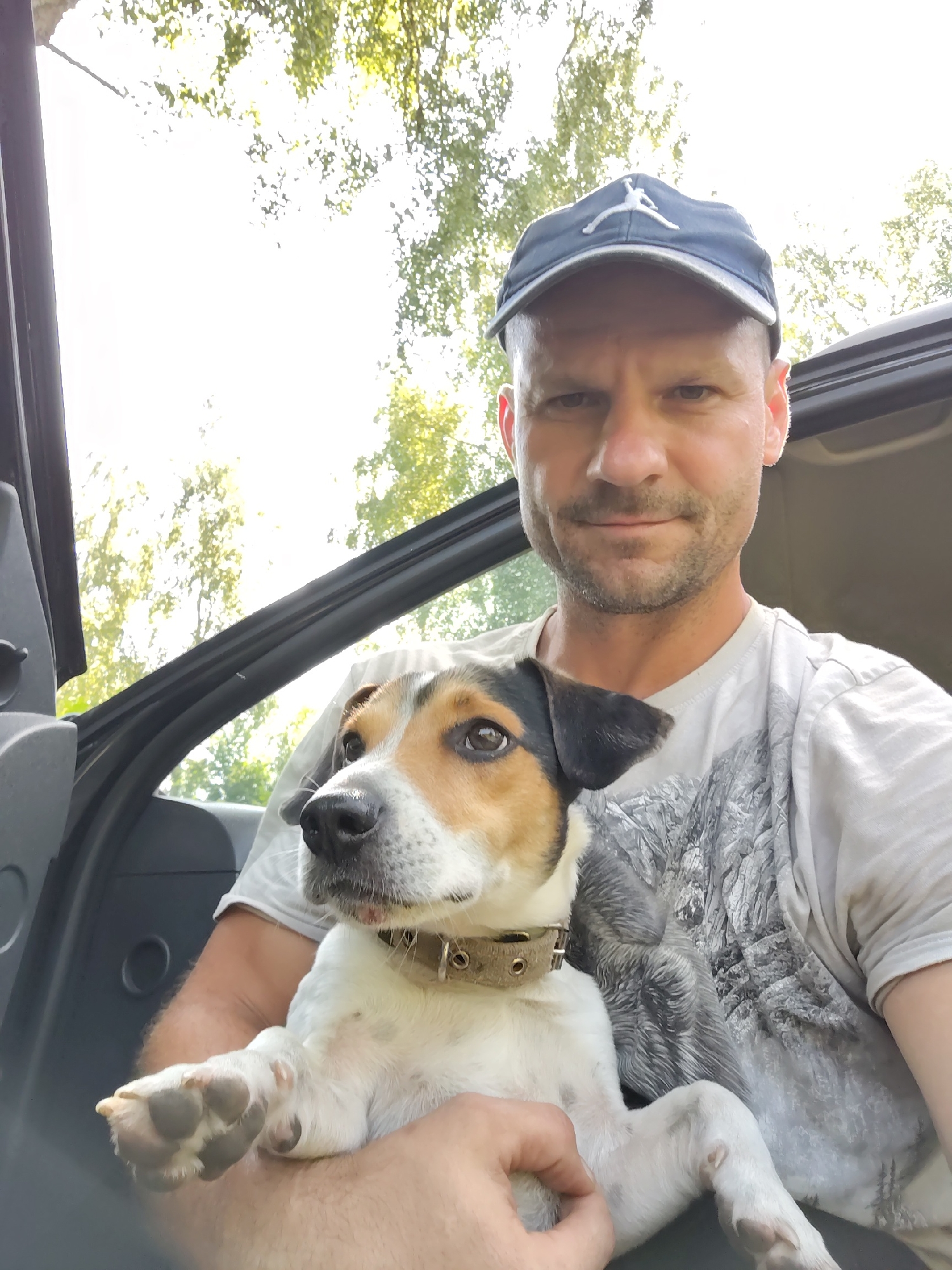A middle-aged Caucasian male with short, light brown hair, stubble, and a light beard is seated in the driver's seat of a vehicle. He is wearing a grey t-shirt with a grey abstract design and a dark blue baseball cap with a white Jumpman logo. He is holding a small terrier mix dog with white, tan, and black markings on its face and body. The dog is wearing a tan collar and is looking towards the camera. The interior of the vehicle, including the door panel, is visible. The background shows bright sunlight filtering through green foliage, suggesting an outdoor setting during the day. The specific location in Чарацянскі сельскі Савет, Belarus, cannot be determined from the visual information provided in the image.