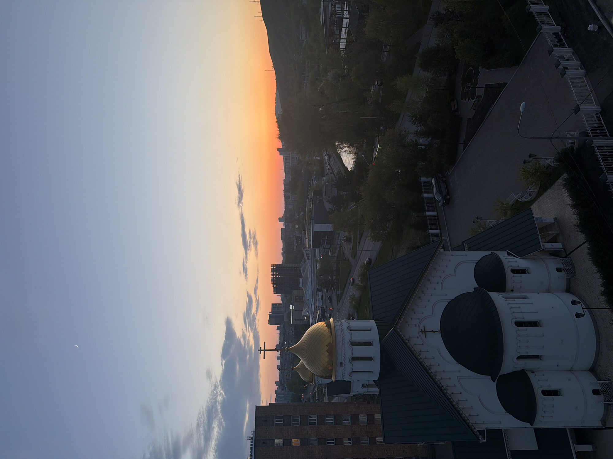Here is a description of the image:
The image depicts a sunset view over a city. The sky transitions from a gradient of orange and red near the horizon to a lighter blue above. A crescent moon is visible in the blue sky. The foreground features a building, likely a church, with a golden dome topped with a cross. The city is densely built, with trees and buildings visible. A hill or mountain range forms the backdrop to the city in the distance. A building with dark green domes and white walls appears in the foreground. The roofs of other buildings are also visible.