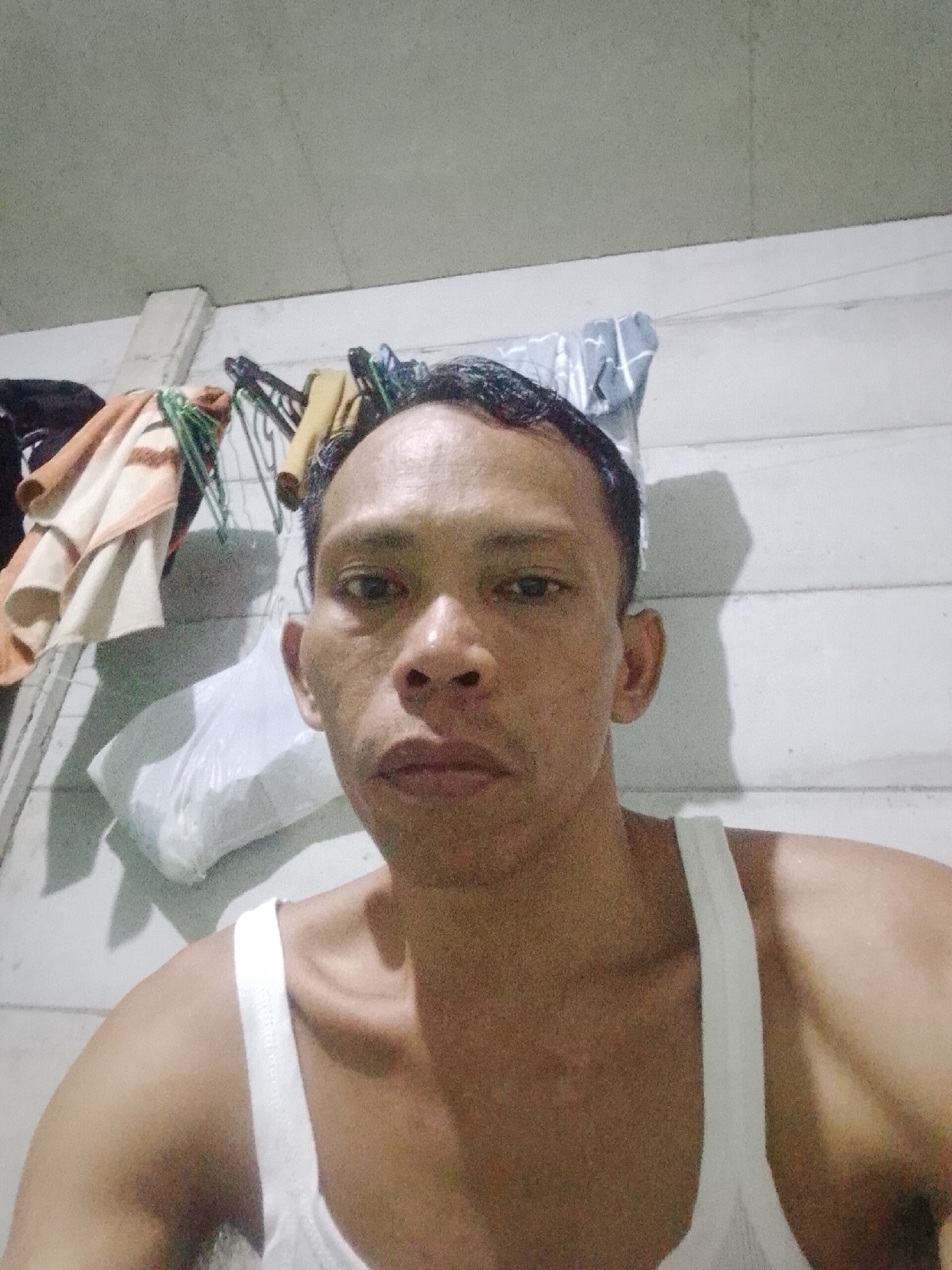 A man with short, dark hair is looking directly at the camera. He has a light-to-medium skin tone, and is wearing a white tank top. He appears to be in a room with white walls and a white ceiling. Behind him are clothes hanging on a rack, including towels, clothes hangers, and a plastic bag. The location is possibly Bukit Ketok, Indonesia.