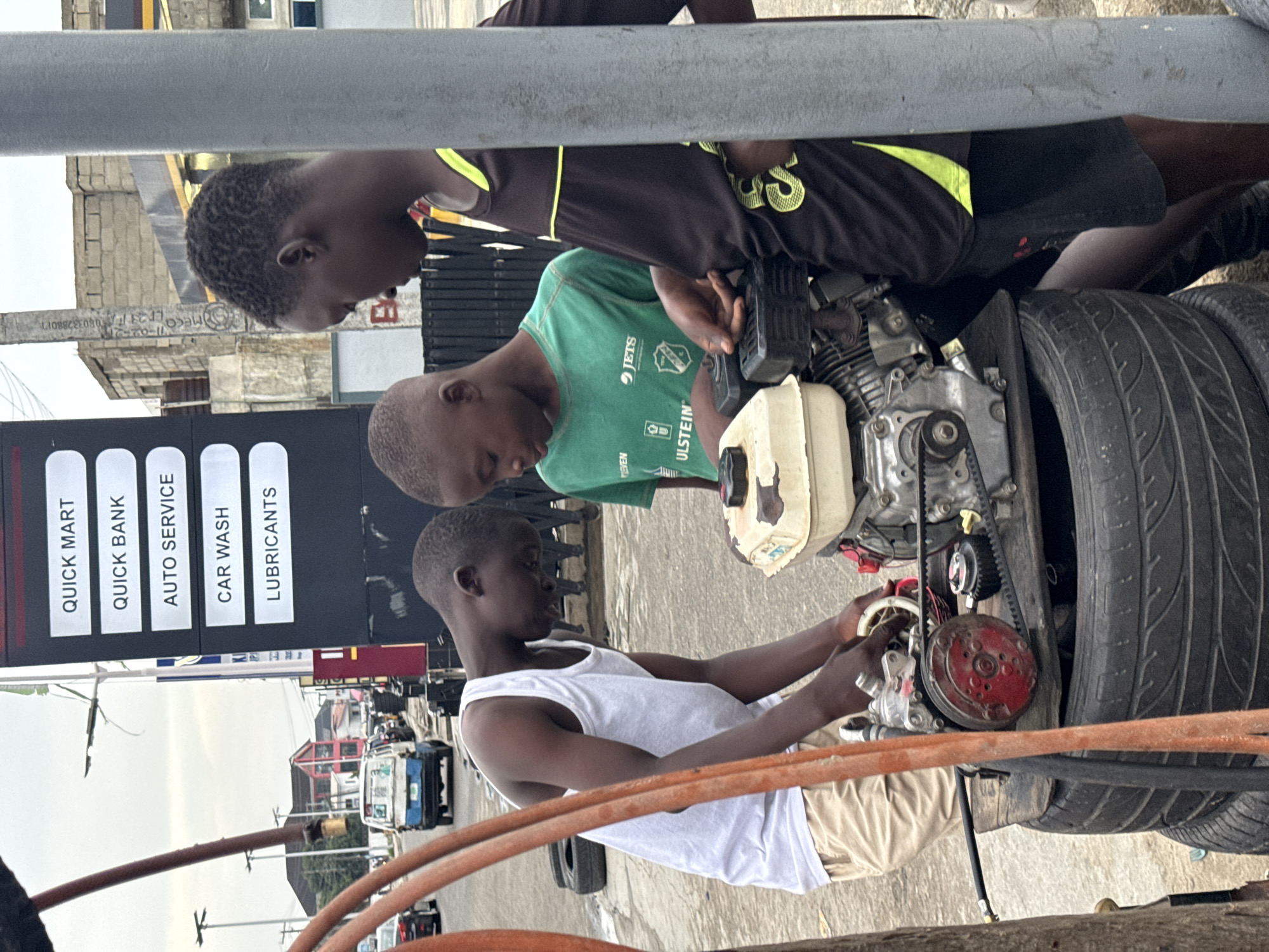 Three boys are gathered around a small engine mounted on a structure seemingly made from tires, working on it. The boy closest to the viewer is wearing a white tank top and tan shorts, focusing on the engine’s red pulley system. Another boy wears a green shirt with logos and text. The third boy is in a black and yellow jersey, peering down at the engine.
Behind them is a black sign advertising "QUICK MART," "QUICK BANK," "AUTO SERVICE," "CAR WASH," and "LUBRICANTS." The backdrop shows a typical city street scene with buildings, vehicles, and power lines, suggesting the location is in Ado, Nigeria.