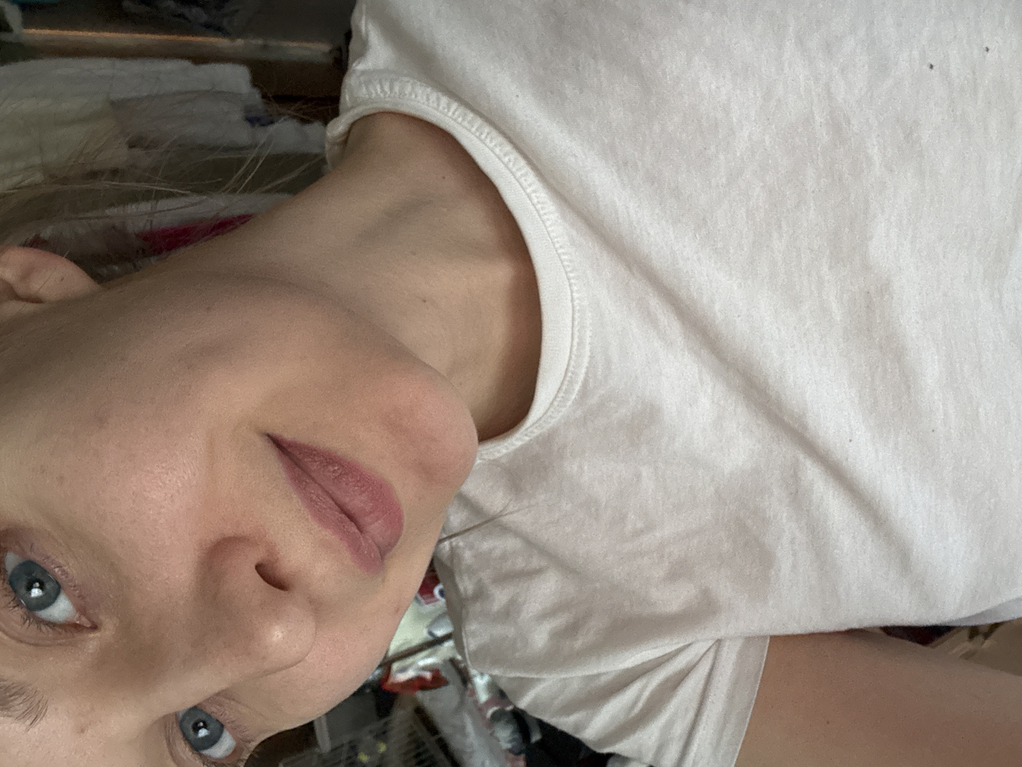 The image is a close-up selfie of a woman with fair skin, blue eyes, and blonde hair, wearing a plain white t-shirt. Her face is slightly angled. The background is cluttered and out of focus, showing various items, possibly clothing, and a metal rack or shelving unit. The location is not discernible from the image and cannot be identified as Izhevsk, Russia, or any other specific location.