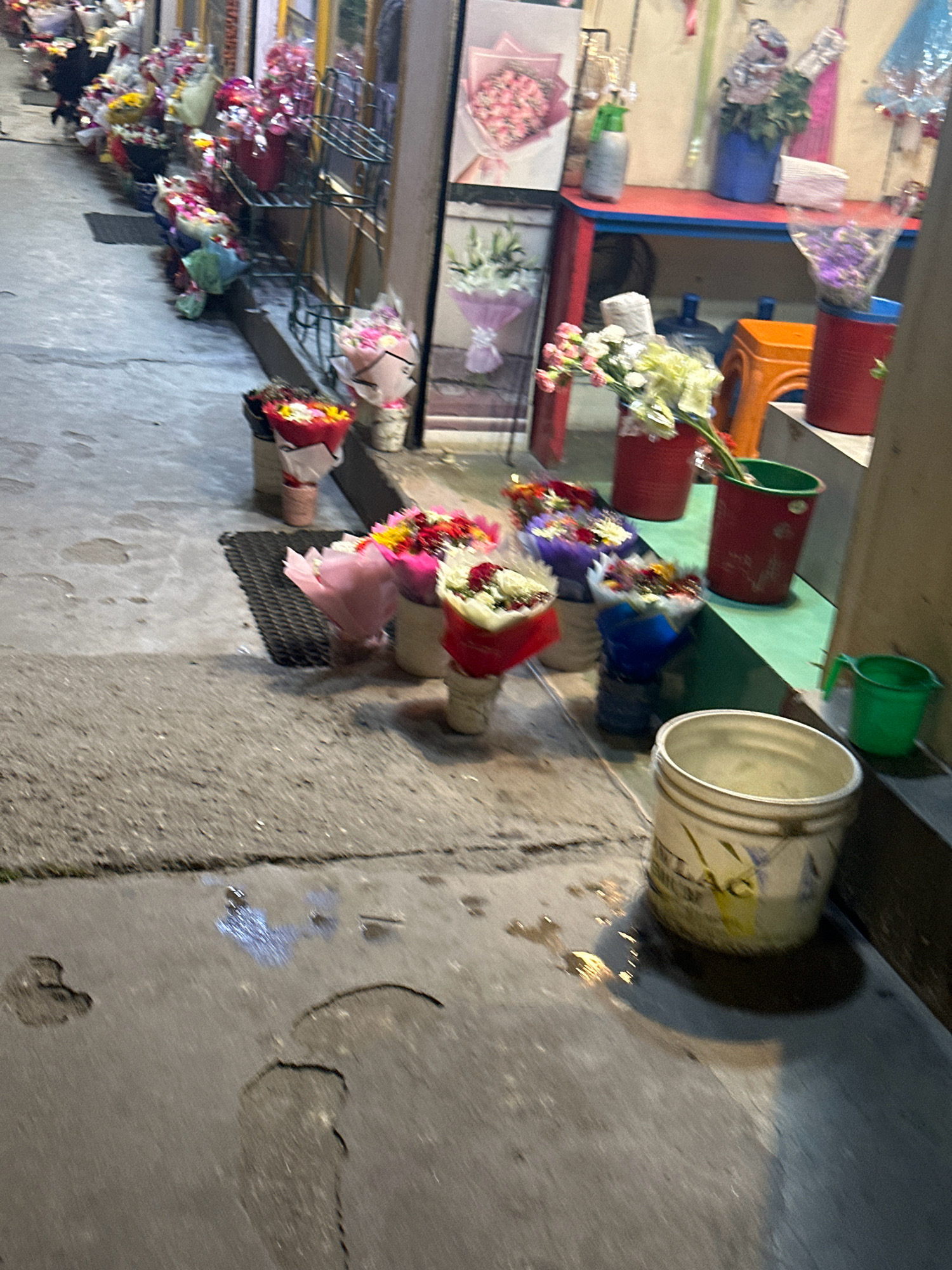 The image depicts a flower stall on a street, likely in Kathmandu Metropolitan City, Nepal, given the visible signage and cultural context. In the foreground, a concrete sidewalk is wet in places, with puddles and an uneven surface suggesting it might have recently rained or been cleaned. A white bucket with some writing on it sits on the edge of a raised platform, casting a shadow. To the right, a small green plastic bucket is visible.
Behind this, a display of flower bouquets is arranged on the ground and on shelves. Several bouquets, wrapped in paper and plastic, are in buckets and containers of various colors, including red and blue. Some of the flowers are pink, white, and red. In the background, shelves with more flowers and plants are visible, along with some plastic containers and what appears to be a red table. The overall impression is of a small, bustling flower shop or stall operating on a street. The lighting suggests it might be evening or early morning.