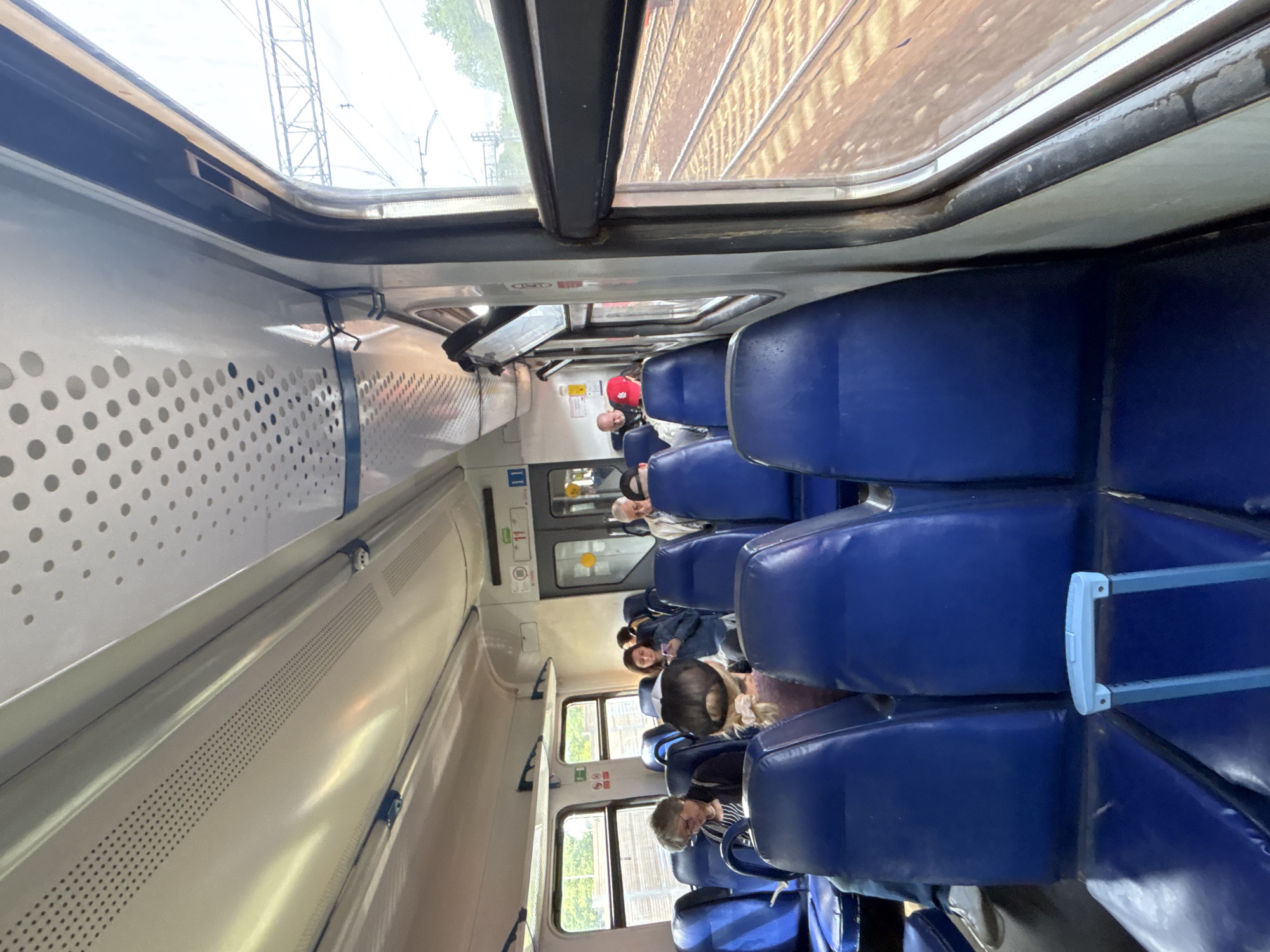 The interior of a train car is visible, likely traveling on a train in Moscow, Russia. The interior is well-lit, and several people are seated in blue upholstered seats with metal frames. Some are looking out of the windows, others seem to be interacting or simply resting. The walls are pale with rows of circular perforations. Through the window, train tracks can be seen.