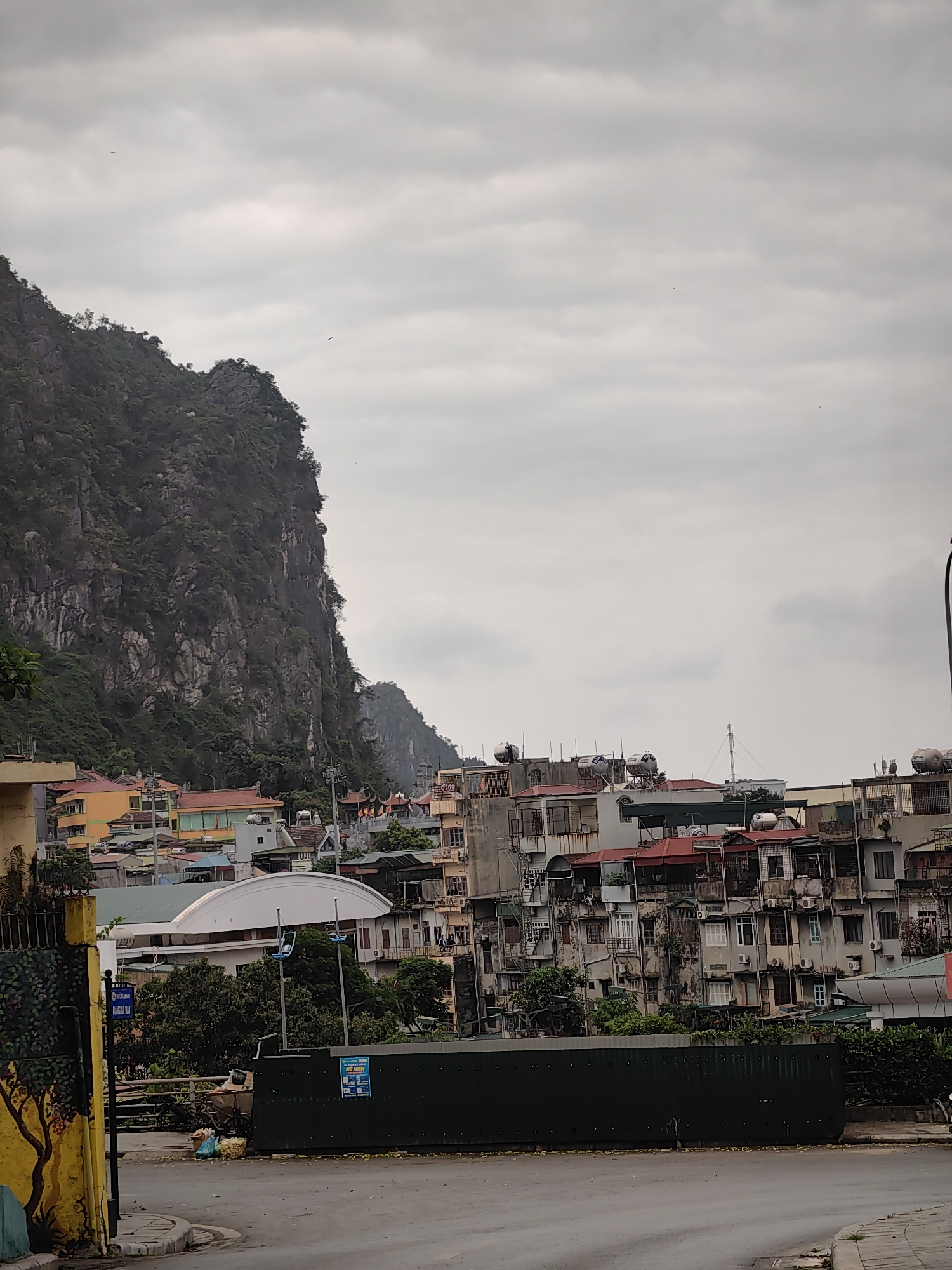 The scene is an overcast cityscape within Hong Gai Ward, Vietnam. A steep, rocky, tree-covered mountain rises abruptly on the left side of the frame, dominating the skyline. To the right of the mountain are dense clusters of buildings, primarily multi-story residential structures, with a mix of architectural styles and colors; many have rooftop water tanks. A low green barrier or fence runs across the midground of the image, partially obscuring the buildings behind. In the foreground, a paved street is visible, damp from recent rain or moisture. A yellow-painted wall with a mural and signage stands on the left edge of the frame.