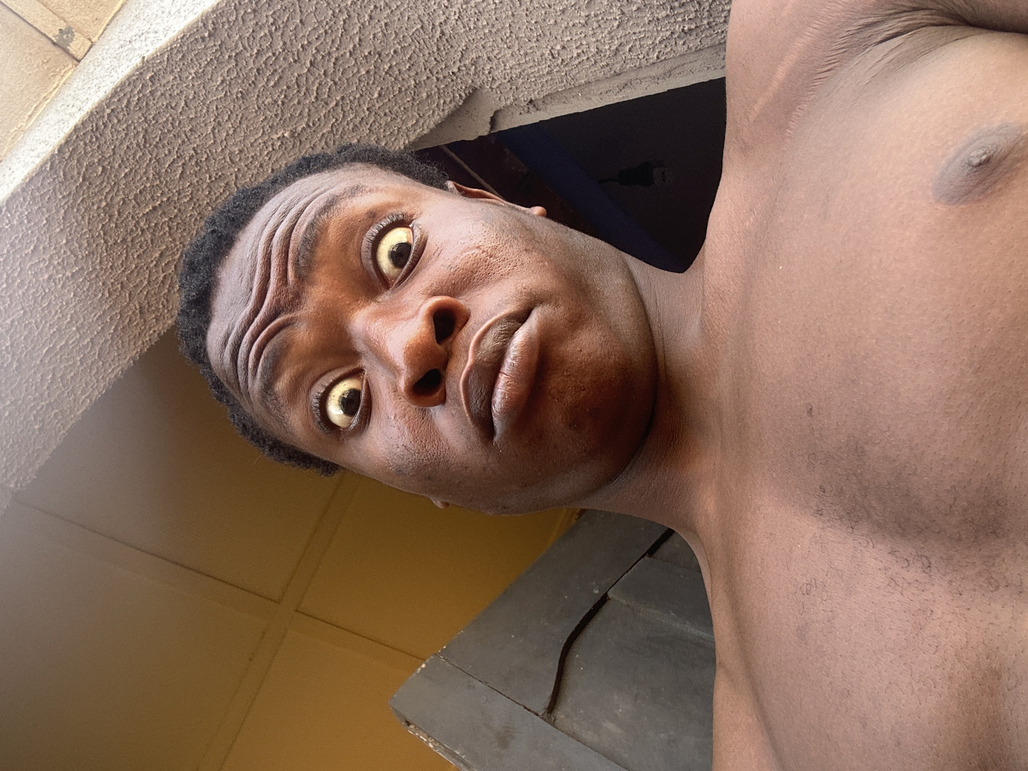 A shirtless man with dark skin and short, curly black hair looks down at the camera with a surprised expression. His eyes are wide and appear slightly gold in color. He is standing in what appears to be a building, with the ceiling visible above him. The walls are painted a light yellow color. The location is Unknown, Unknown.