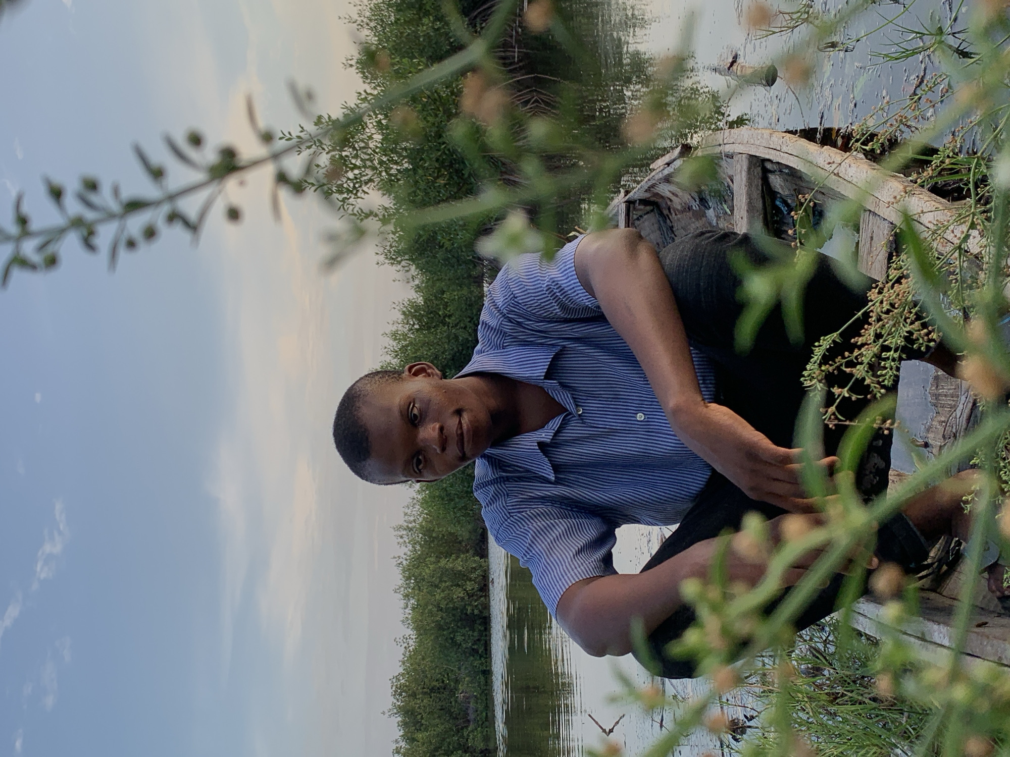 A young man of African descent, with short dark hair and wearing a blue and white striped button-up shirt and dark trousers, is pictured crouching inside a rustic wooden boat on calm water. He is looking directly at the camera with a gentle smile, appearing relaxed and friendly.
The setting is an outdoor, natural environment, likely a river or lagoon, with dense green vegetation, possibly mangroves, lining the water's edge in the background. The vegetation is reflected in the still surface of the water, which also shows reflections of the soft blue sky with a few faint clouds. The lighting suggests either late afternoon or early morning.
In the foreground, out-of-focus green foliage with small buds or berries partially frames the image, adding depth and a natural element to the composition. The boat appears to be a simple, weathered wooden vessel, suggesting its use for local transport or fishing. The scene conveys a peaceful atmosphere, capturing a moment of daily life in a natural, waterside community. There is no visible text in the image. The details are consistent with the given location context of Ibasa, Nigeria.