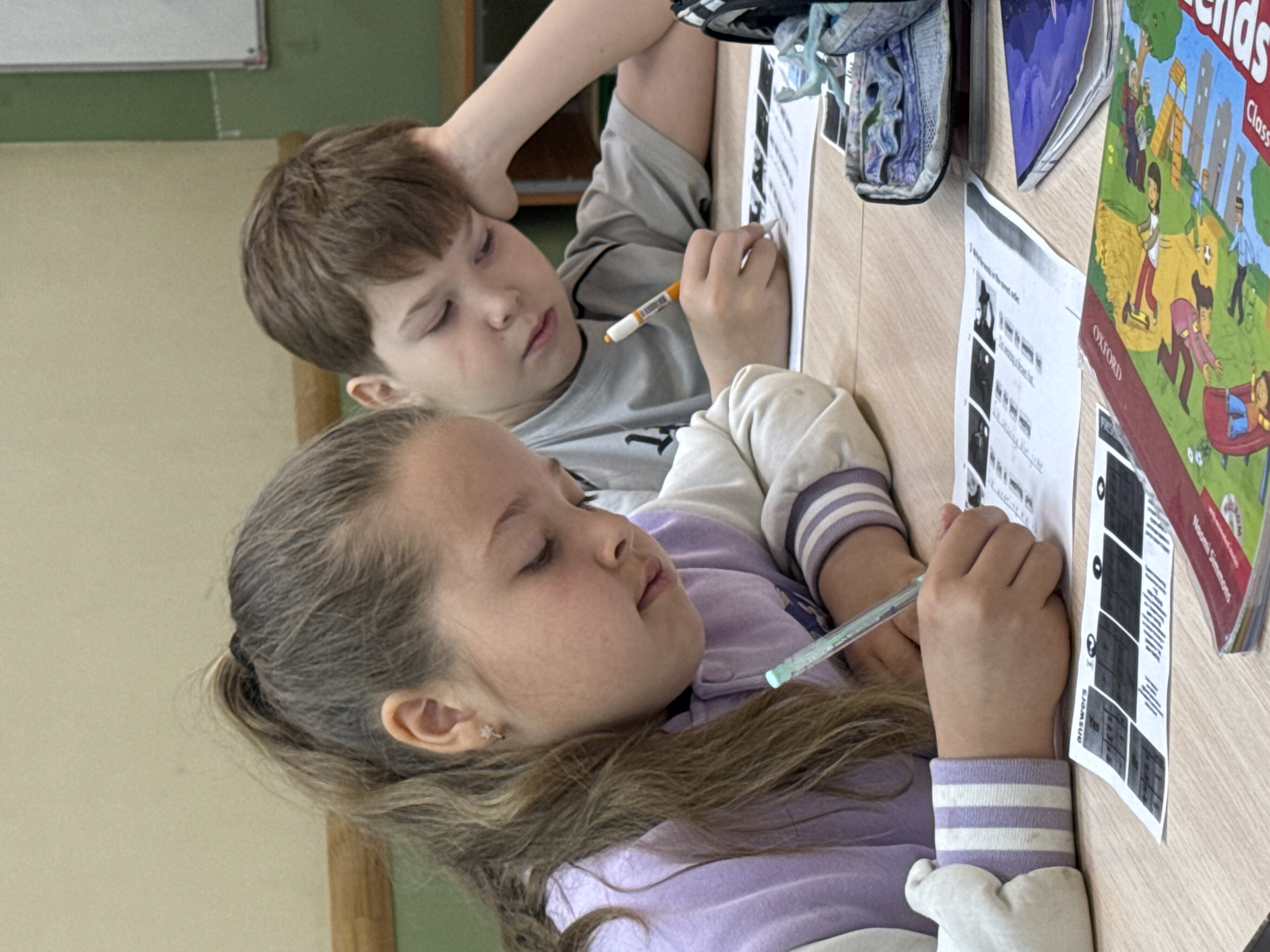 Two children, a girl and a boy, are seated at a wooden table. The girl, positioned closer to the camera, has light brown hair pulled back in a ponytail and wears a light purple and white striped jacket. She holds a pen in her right hand and looks at the worksheet in front of her. The boy behind her has short brown hair and a gray shirt. He also holds a pen and appears to be working. On the table, there are worksheets with text, a textbook titled "Oxford Friends," and a pair of blue and white sneakers. The background shows a light-colored wall. The location is presumed to be Perm, Russia, based on external information.