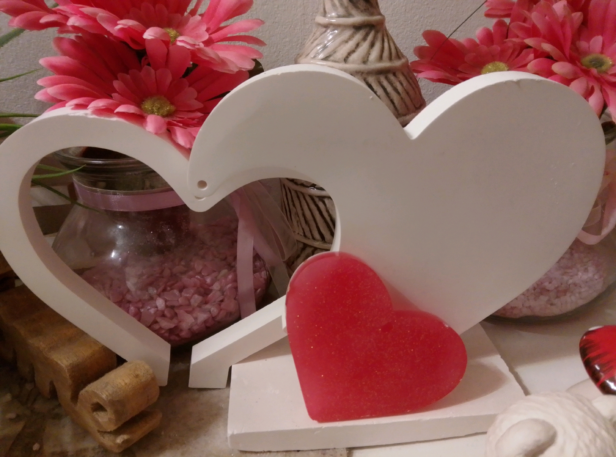 The image depicts a still life arrangement of decorative items. In the foreground, a large, white, heart-shaped cutout is prominently displayed. A smaller, bright red, translucent heart, possibly made of resin or glass, rests on a white rectangular base in front of the larger heart. Behind the white heart, a glass jar filled with small, pink, crystalline objects is visible, tied with a pale pink ribbon. Further in the background, a decorative ceramic vase with a textured, striped pattern stands amongst a cluster of vibrant pink artificial flowers. The overall setting appears to be indoors, with a neutral-colored wall as the backdrop and a textured surface that could be a table or shelf beneath the objects. The lighting suggests a soft, ambient light source, casting subtle shadows. There are no people or animals present in the image, and no specific location in Wedel, Germany is indicated or discernible.
