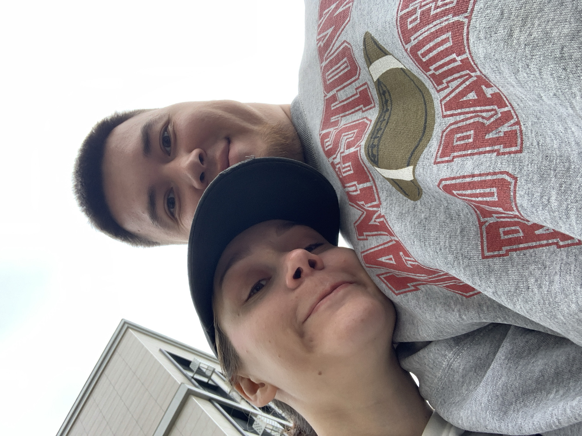 A close-up, low-angle, selfie-style photograph shows two individuals, presumably a couple, outdoors. The person on the left, a female wearing a black baseball cap and leaning her head on the shoulder of the person on the right, is looking towards the camera with a slight smile. The person on the right, a male, is smiling and looking directly at the camera. Both are wearing a grey sweatshirt with red lettering and an image of a football. The background consists of a building with angular architecture and a bright, overcast sky. There is no indication of the city of Stavropol, Russia, in this image.