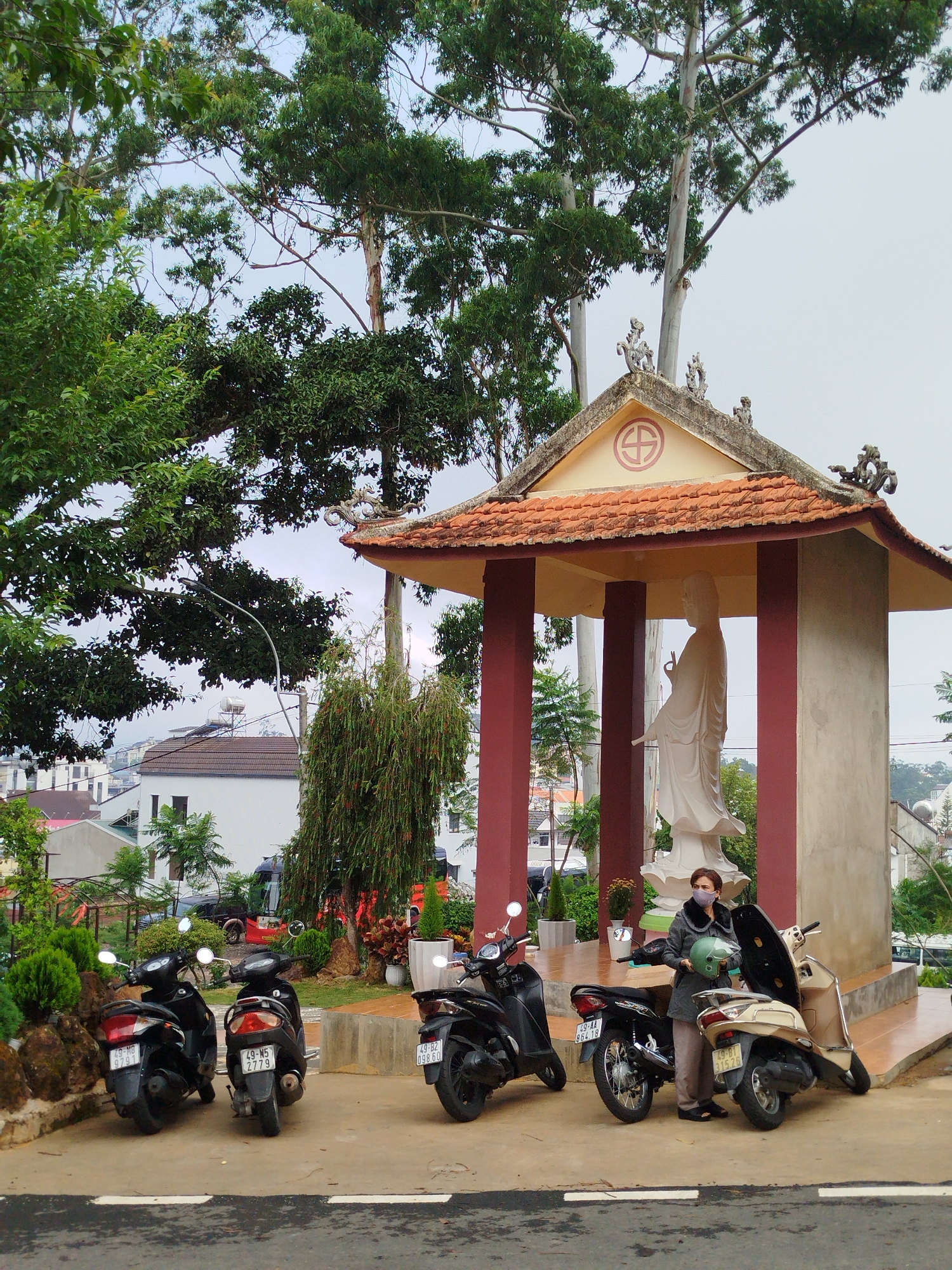The image depicts a scene in Da Lat, Vietnam. In the foreground, there are five parked motorbikes along the edge of a road. A woman wearing a grey coat and a face mask stands next to one of the motorbikes, holding a green helmet. Behind the motorbikes, there is a raised concrete platform with a small open-air temple structure. The temple has a red tile roof, beige walls, and red columns. Inside the temple stands a white statue. The background features a residential area with houses and lush trees. The sky appears overcast.
