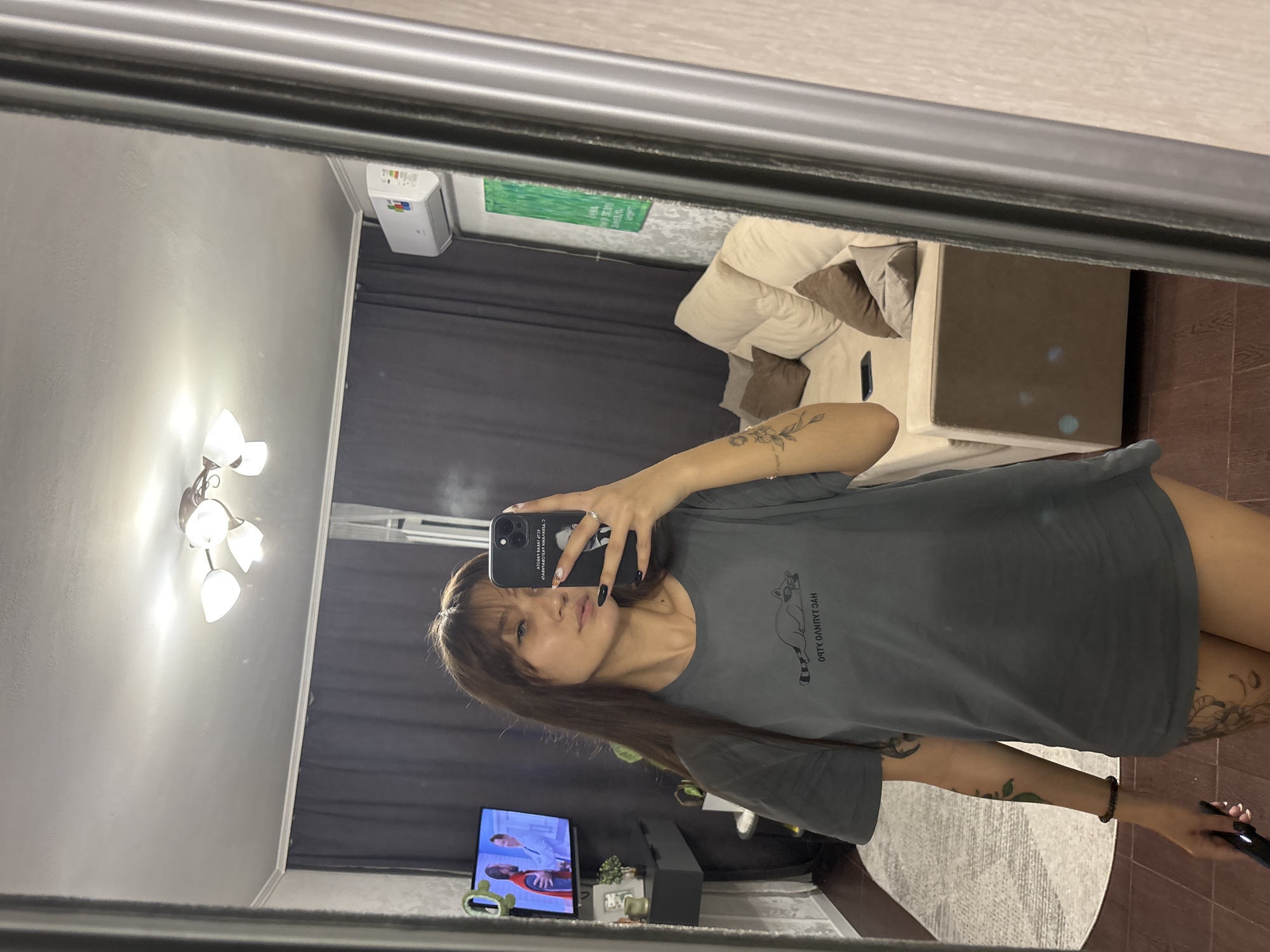 The media file shows a young woman taking a mirror selfie in what appears to be a bedroom or living area in Novokuznetsk, Russia. The woman has long, dark hair and is wearing a dark grey t-shirt with a graphic of a raccoon and some text in a foreign language. She has manicured nails and several tattoos visible on her arms.
In the background, reflected in the mirror, is a couch with several pillows, a television displaying a program with two people, and a lamp with three lights on the wall. There is also a white unit mounted on the wall, possibly an air conditioner or heater. The overall impression is that of a casual, indoor setting. The lighting suggests it could be daytime, or the room is well-lit. There are no explicit indicators of the weather or time of day beyond the indoor setting.
The text on the t-shirt appears to be in Cyrillic script and reads "ОЯУК ОЛЯПЯГ ЗАЯ". The screen of the phone she is holding also displays text, but it is not clearly legible. The woman's expression is neutral, and she is looking directly into the camera.