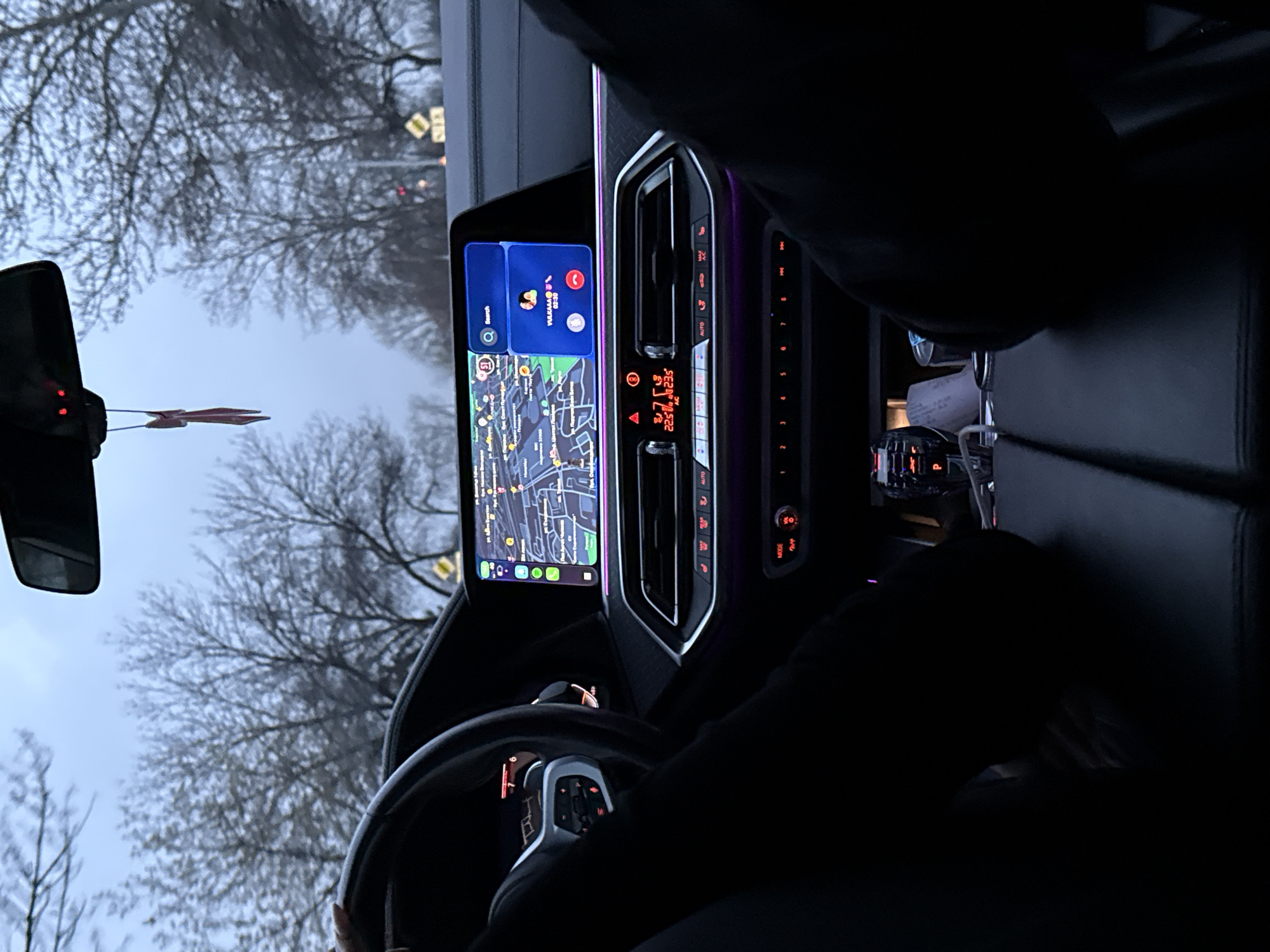 The image is taken from the interior of a BMW car, showing the dashboard, steering wheel, and the road ahead through the windshield. The car's infotainment system is displaying a map, showing it is located in Plovdiv, Bulgaria, specifically near ул. Цар Асен, ул. Княз Александър Батенберг, and ул. Съборна. The climate control panel indicates a temperature of 22.5 degrees Celsius. There is a rear-view mirror and a car air freshener hanging from it. The outside environment shows trees and a cloudy sky. A driver is present, their hands are on the steering wheel. The back seats are dark.